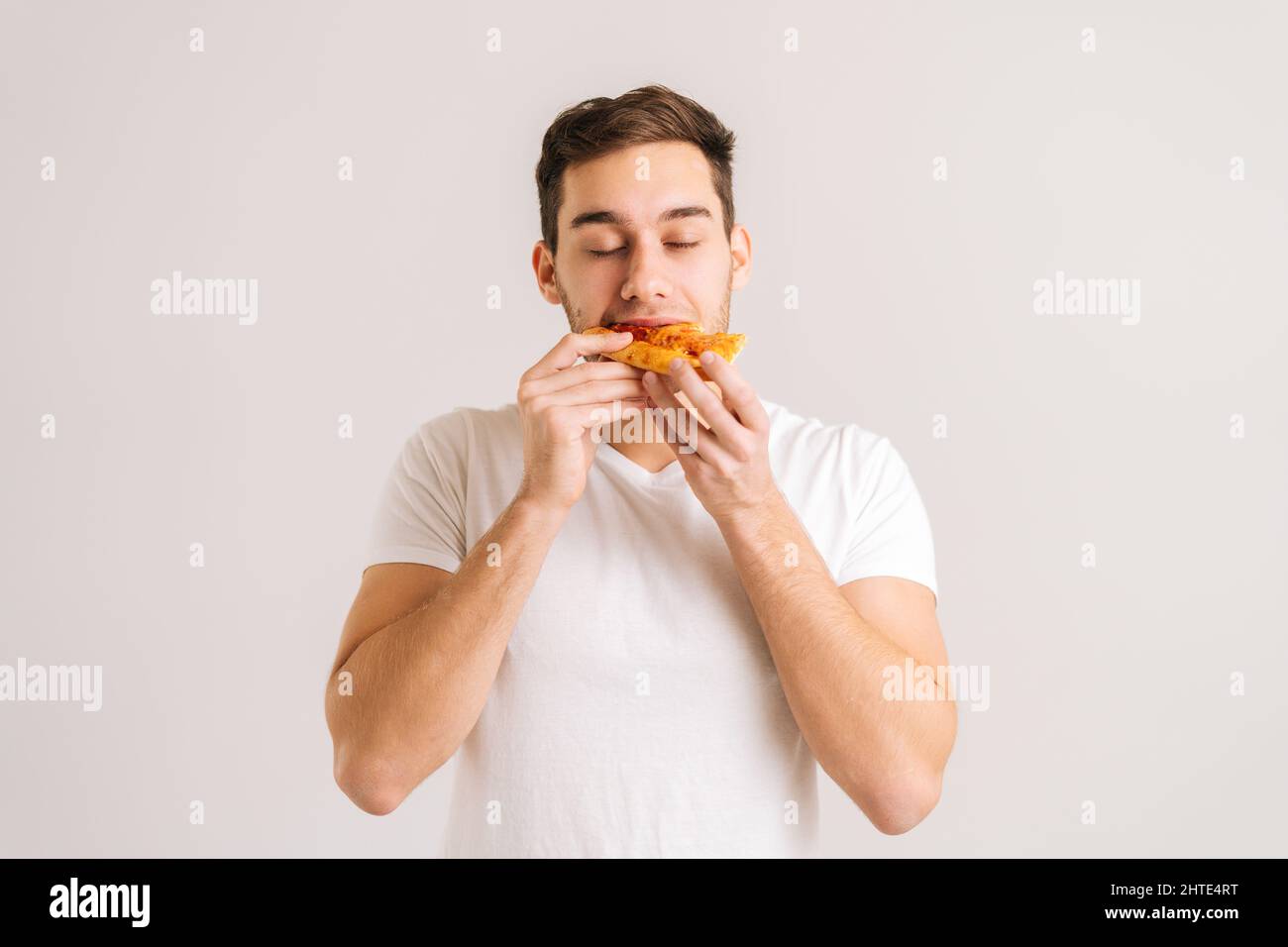 Portrait of hungry young man with enjoying bitting delicious slice of pizza, with closed eyes from pleasure on white isolated background. Stock Photo