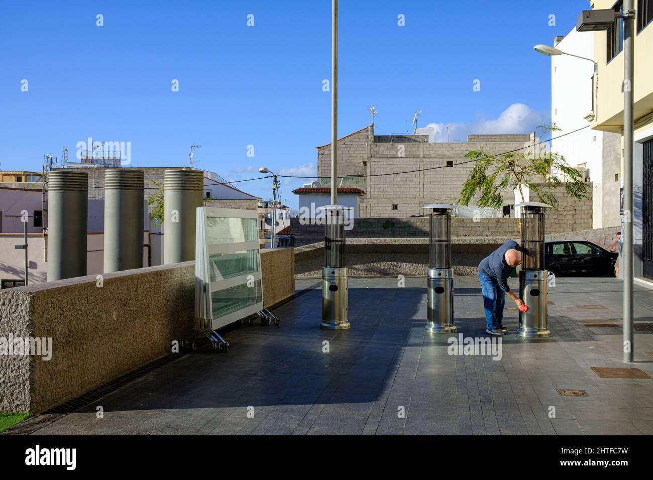 Man cleaning patio heaters on a terrace mirrored by the ventilation shafts from underground parking in the plaza of Playa San Juan, Stock Photo