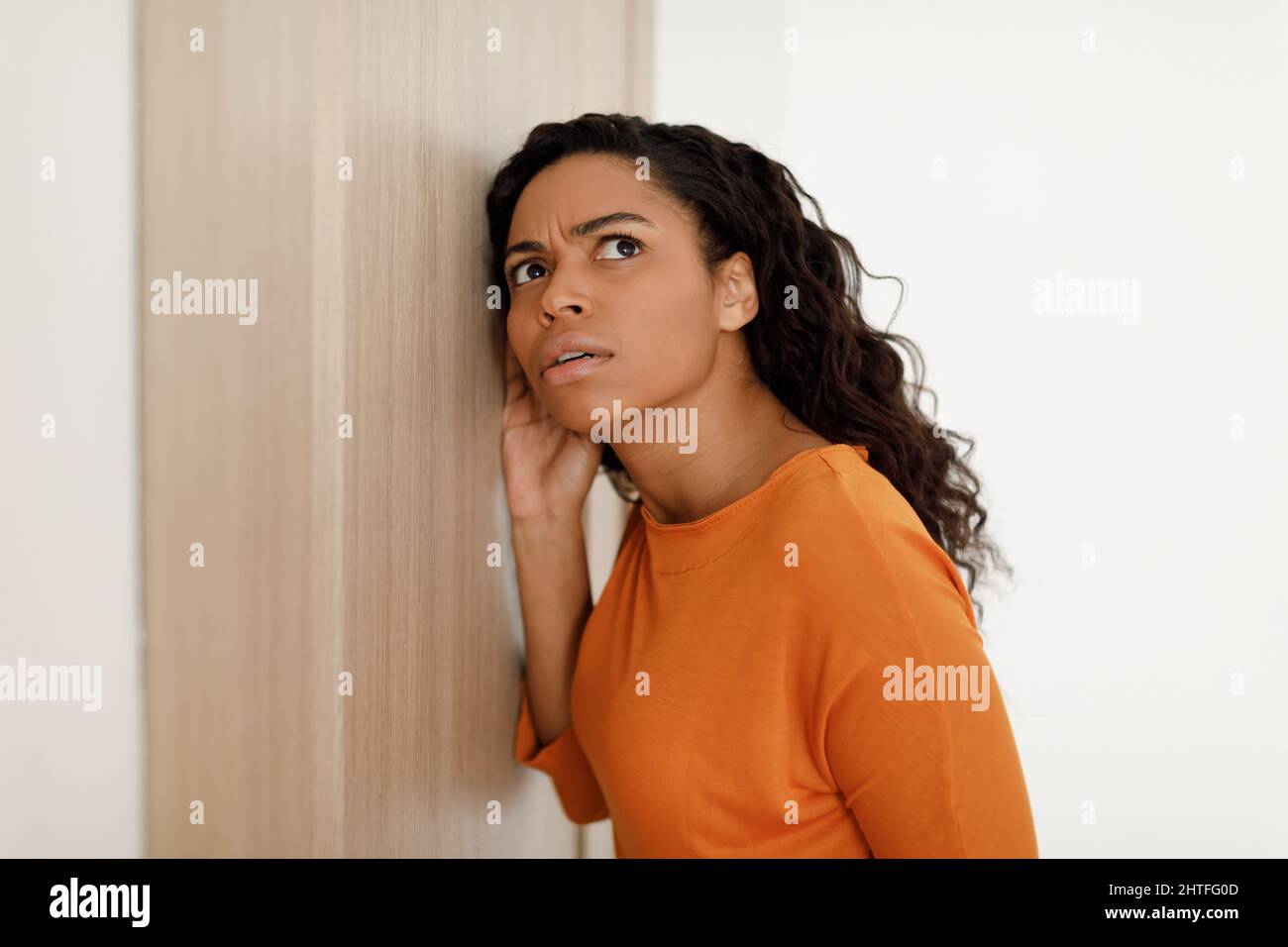 Worried African American Woman Listening Through Closed Door Standing Indoor Stock Photo
