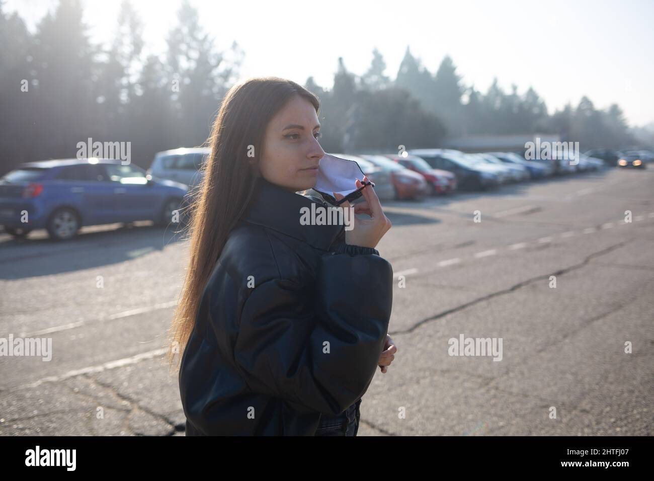 Woman wearing a mask, black jacket and jeans walking in a parking area Stock Photo