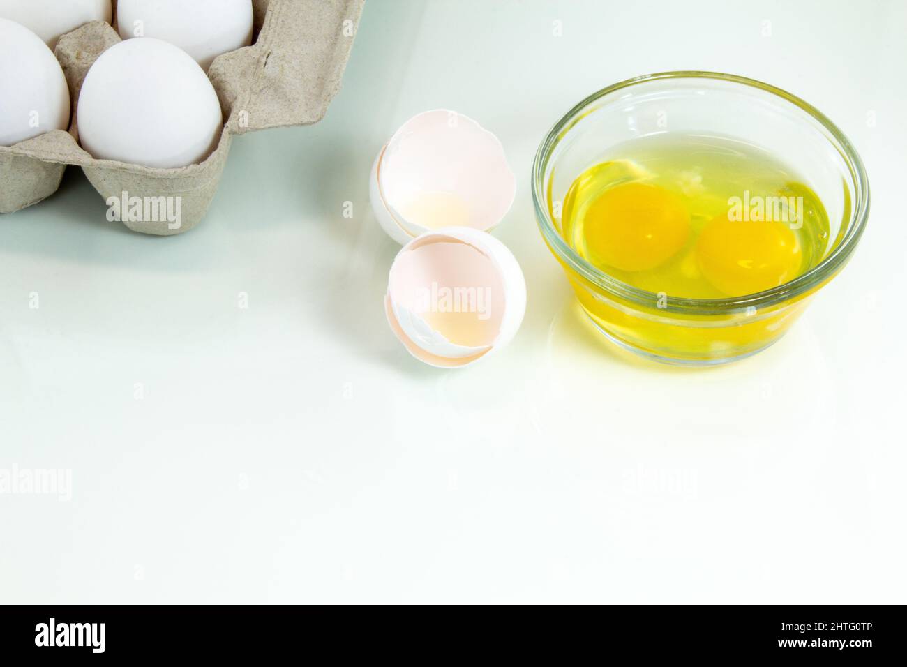 Two broken eggs in a glass bowl with a carton of whole white eggs. White background. Stock Photo