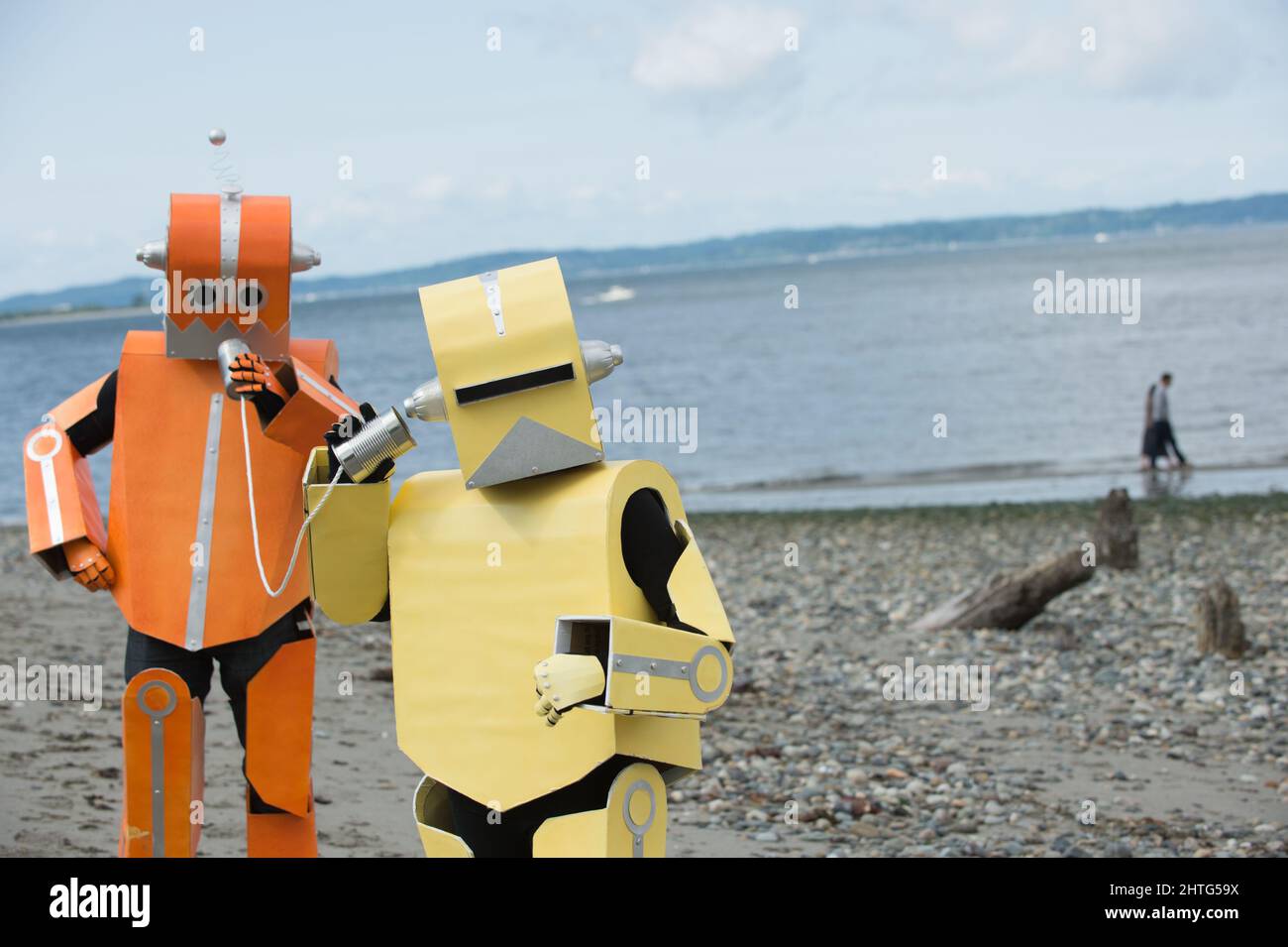 Two colorful robots communicating with each other through tin can telephone while on the beach. People walk by in a distance while robots talk. Stock Photo