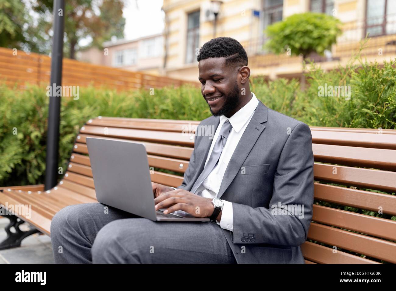 Remote work outdoors. Confident african american businessman or manager in suit using laptop, sitting on bench outside Stock Photo