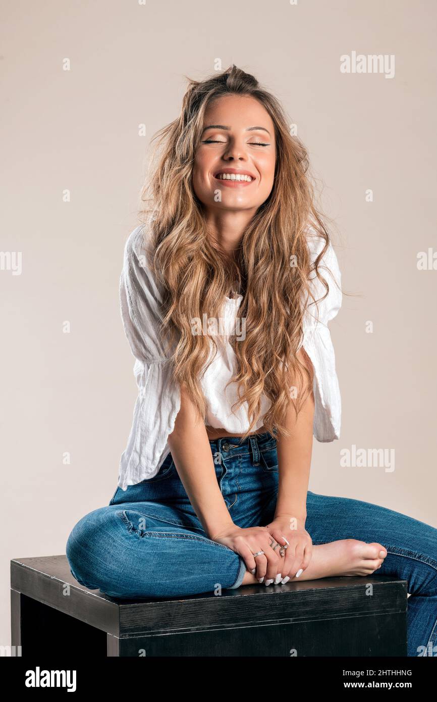 Cheerful blond lady with long wavy hair dreaming with closed eyes while sitting in studio against gray background Stock Photo