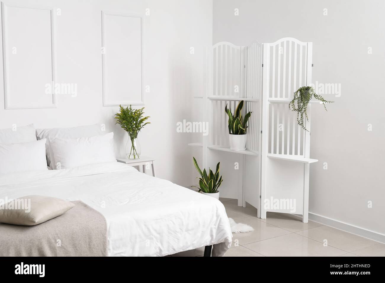 Comfortable bed and folding screen with houseplants near white wall