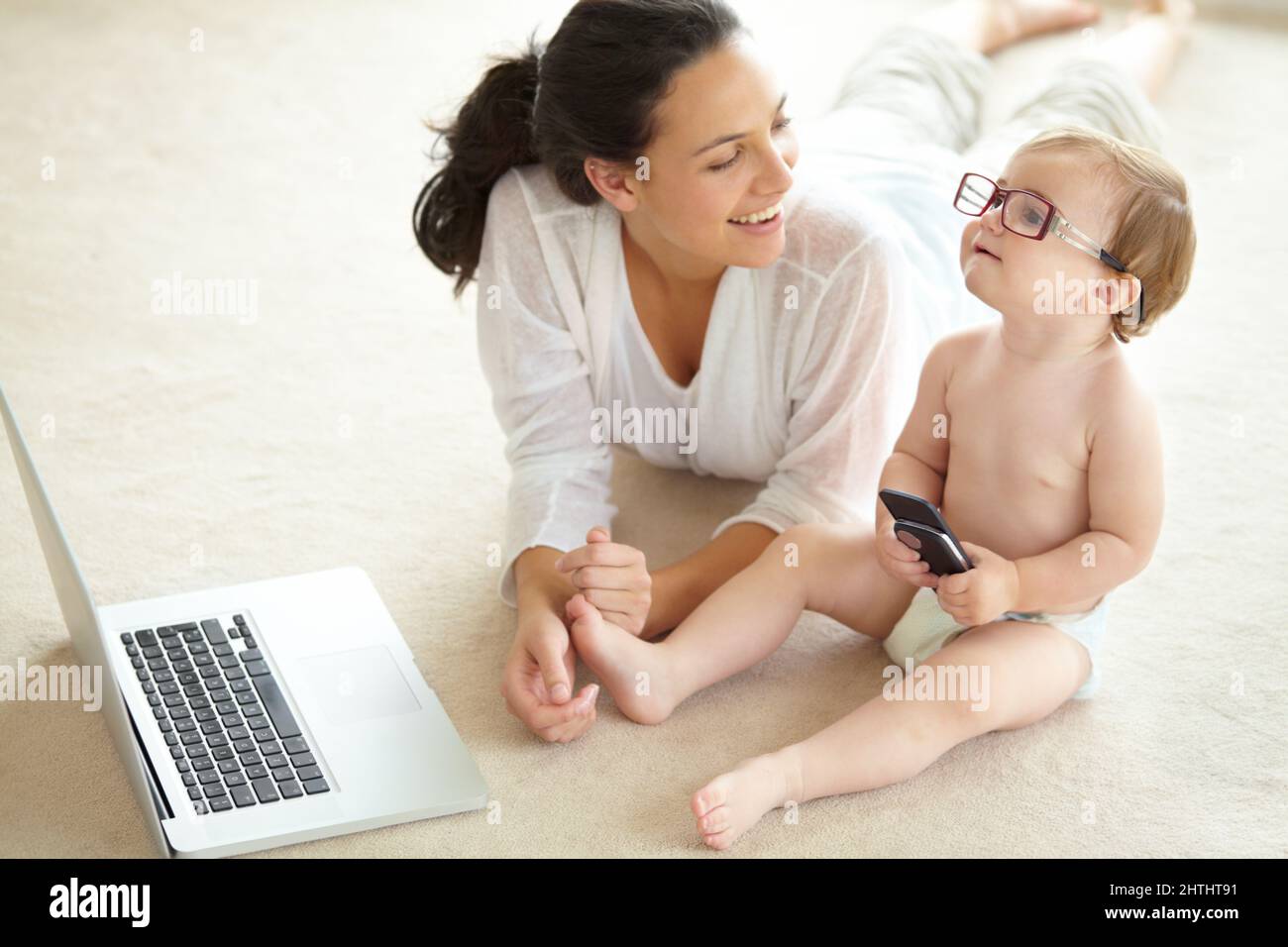 Shes destined for great things. A cute little baby wearing her mothers glasses and holding a cellphone while sitting in front of a laptop. Stock Photo