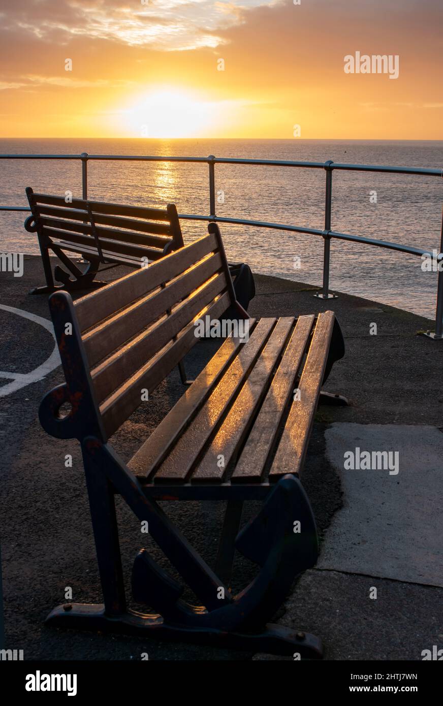 Empty seaside benches face out to sea, bathed in the glow of a warm golden sunrise. Good symmetry and amber and orange light combine with shadows Stock Photo
