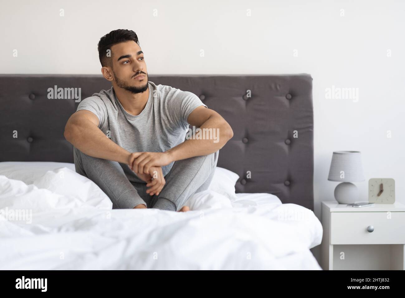 Portrait Of Thoughtful Young Arab Man Sitting On Bed And Looking Away Stock Photo
