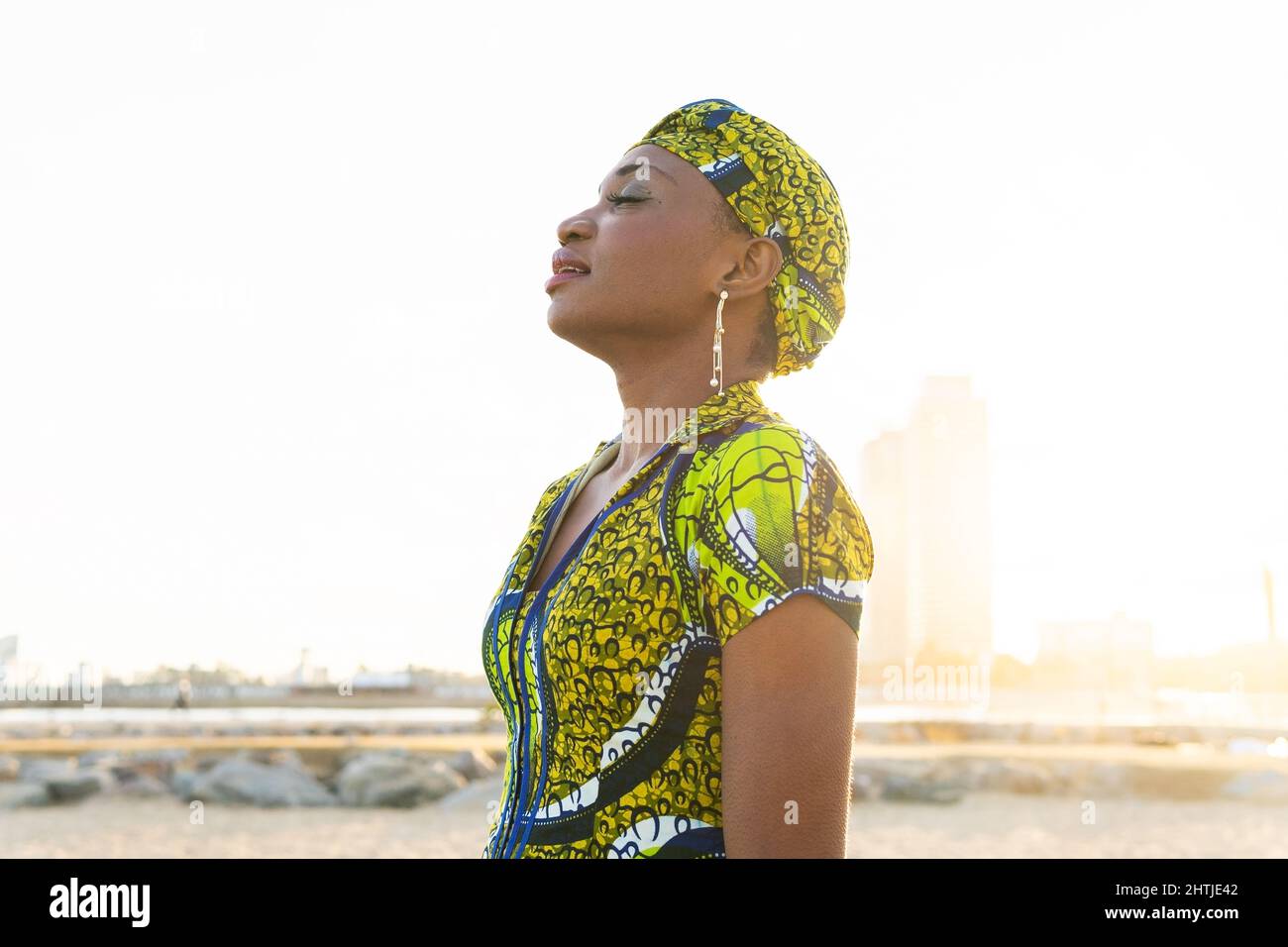 Relaxed African Woman in traditional headscarf standing on sandy beach and meditating at sundown Stock Photo