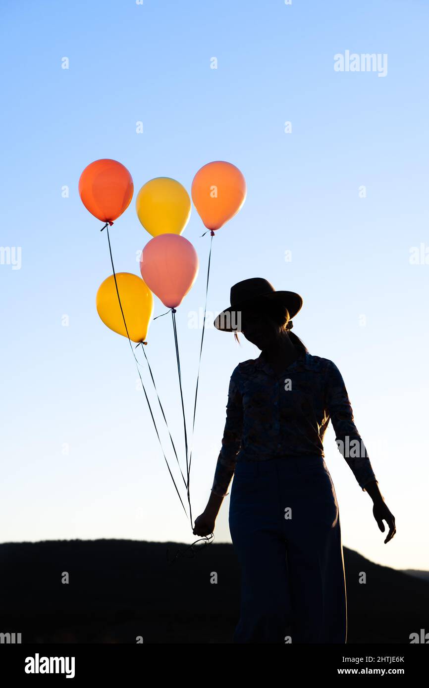 Silhouette of unrecognizable female in hat standing with heap of colorful balloons against cloudless sky at sunset time against mountain ridge Stock Photo