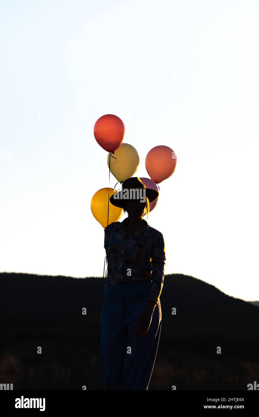 Silhouette of unrecognizable female in hat standing with heap of colorful balloons against cloudless sky at sunset time against mountain ridge Stock Photo