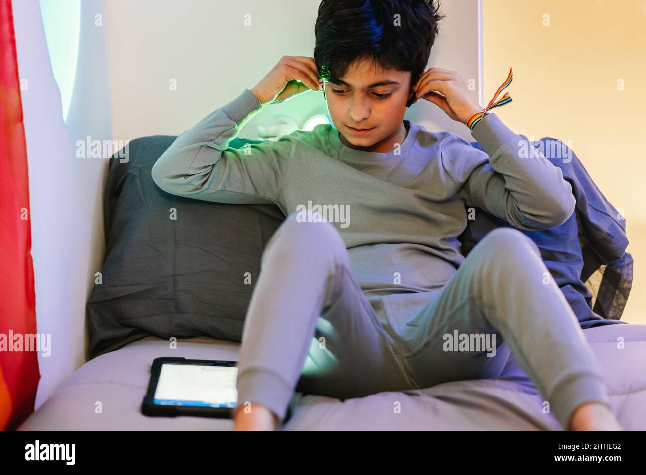 Arab teen boy with rainbow bracelet on wrist wearing pajama adjusting wireless earphones while sitting on bed and using tablet Stock Photo
