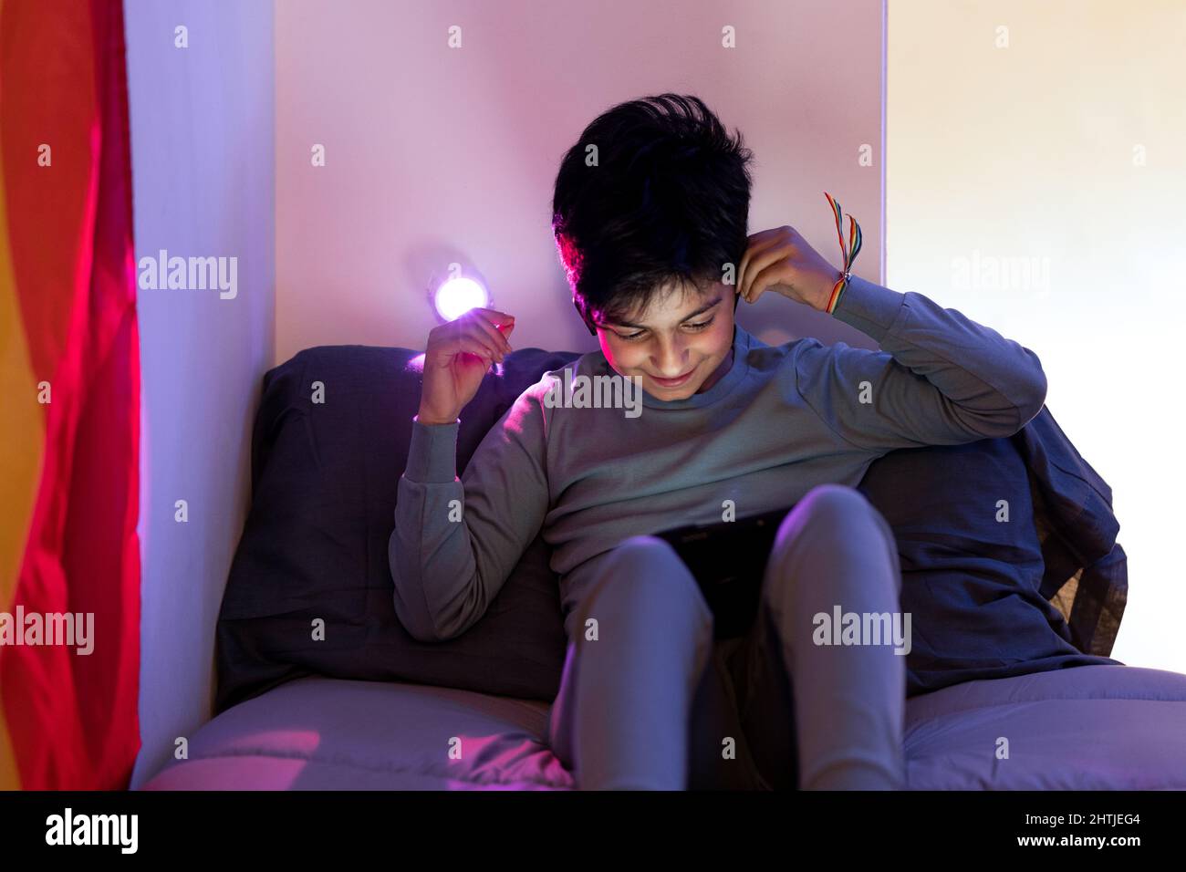 Happy Arab teen boy with rainbow bracelet on wrist wearing pajama adjusting wireless earphones while sitting on bed and using tablet Stock Photo