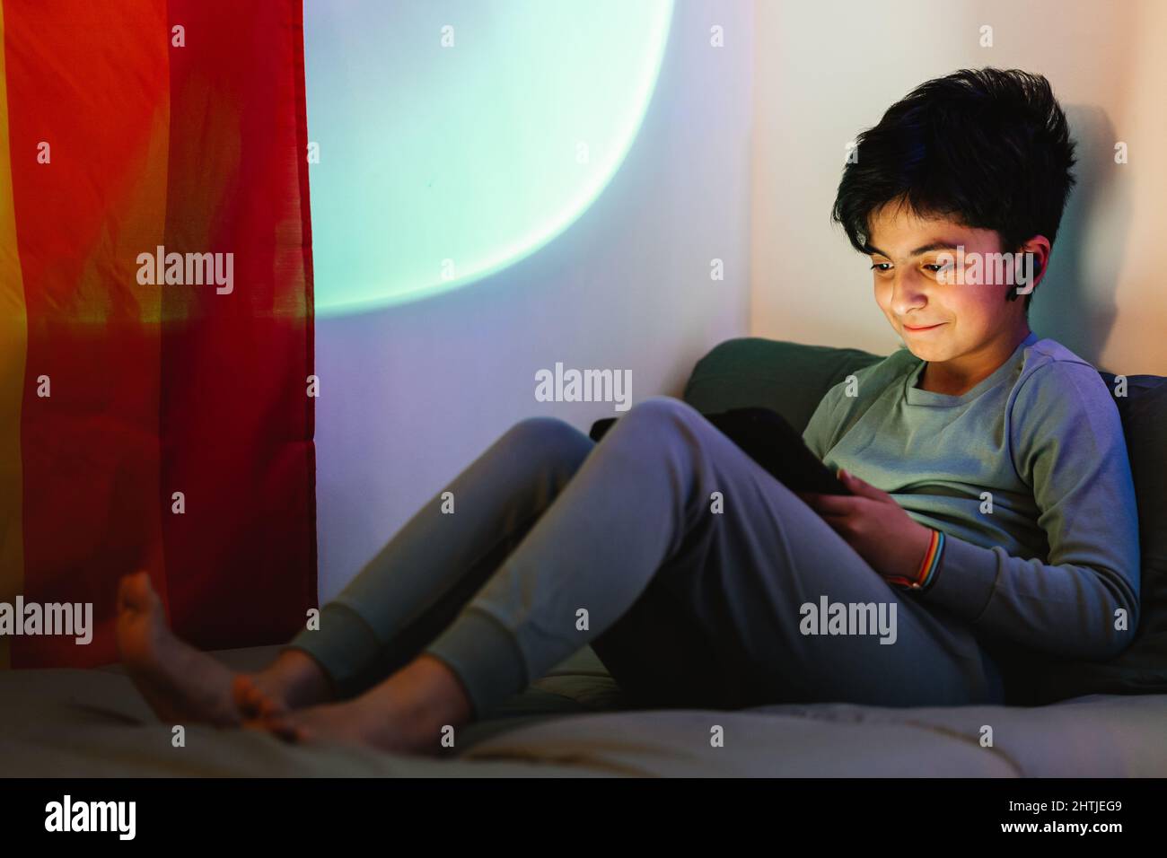 Arab happy teen boy with rainbow bracelet on wrist wearing pajama with wireless earphones while sitting on bed and using tablet Stock Photo