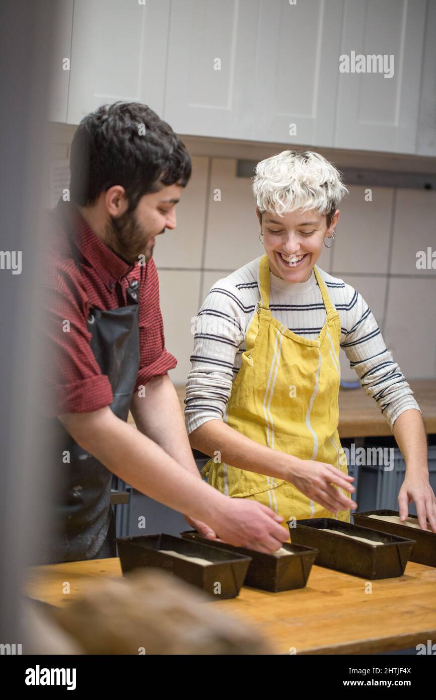 Cheerful couple of bakers in aprons putting bread dough into metal baking pan while working together on kitchen of bakery Stock Photo