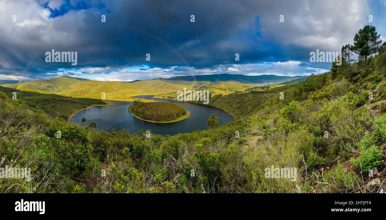 Breathtaking scenery of rainbow over calm river flowing through hilly terrain against cloudy blue sky in Meandro Del Melero National Park in Spain Stock Photo