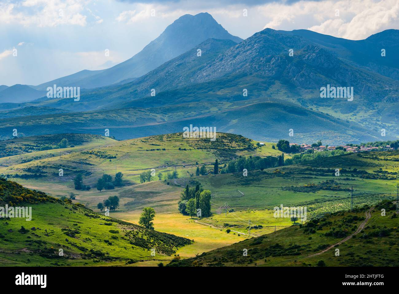 Spectacular scenery of small settlement located on grassy hill slope near massive mountain range against cloudy sky in sunlight Stock Photo
