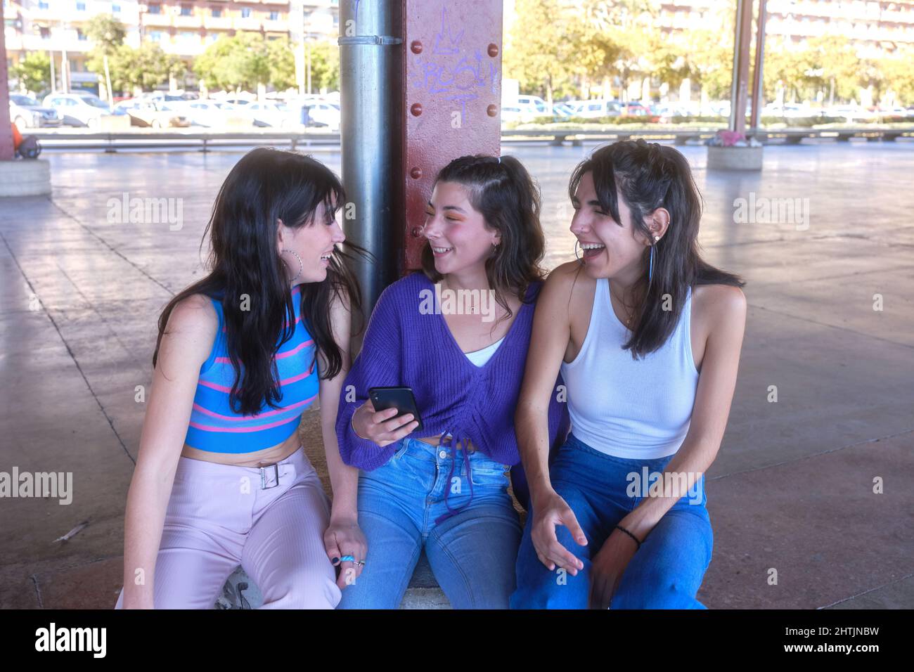 Three friends laughing while using the mobile sitting on an urban area Stock Photo