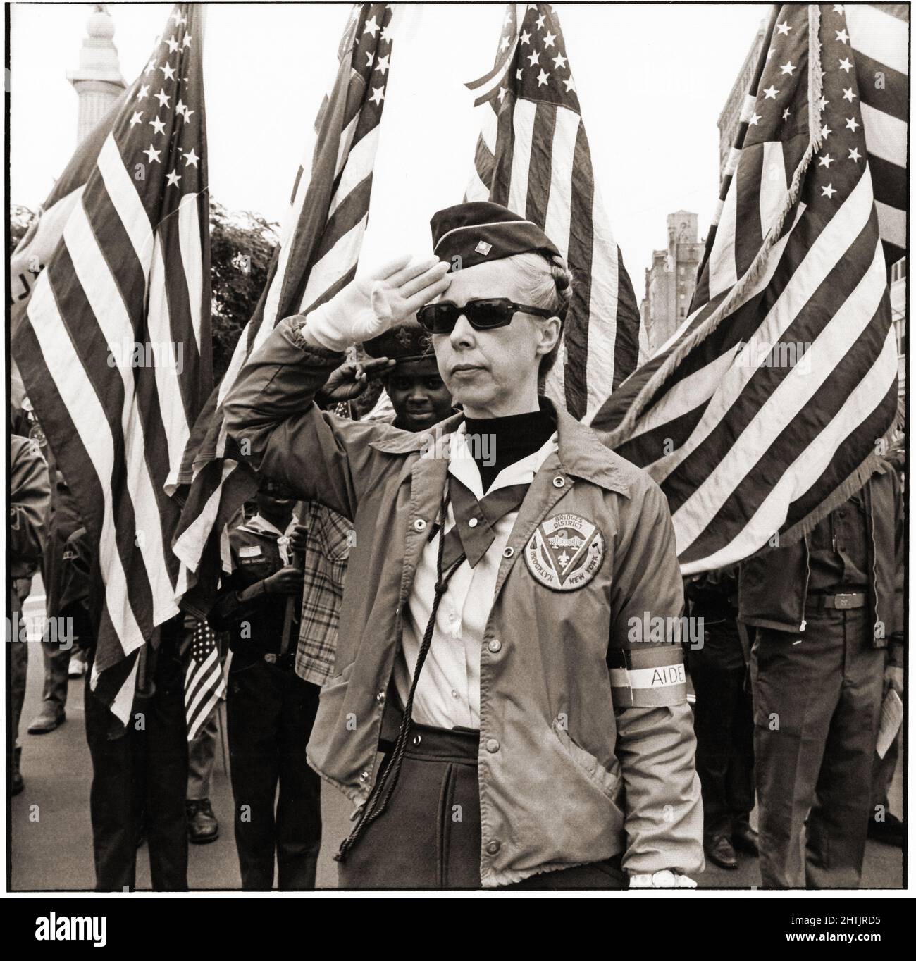 A cub scout leader salutes at the playing of the National Anthem. Behind her are scouts holding American flags. At Grand Army Plaza in Brooklyn, New York circa 1977. Stock Photo