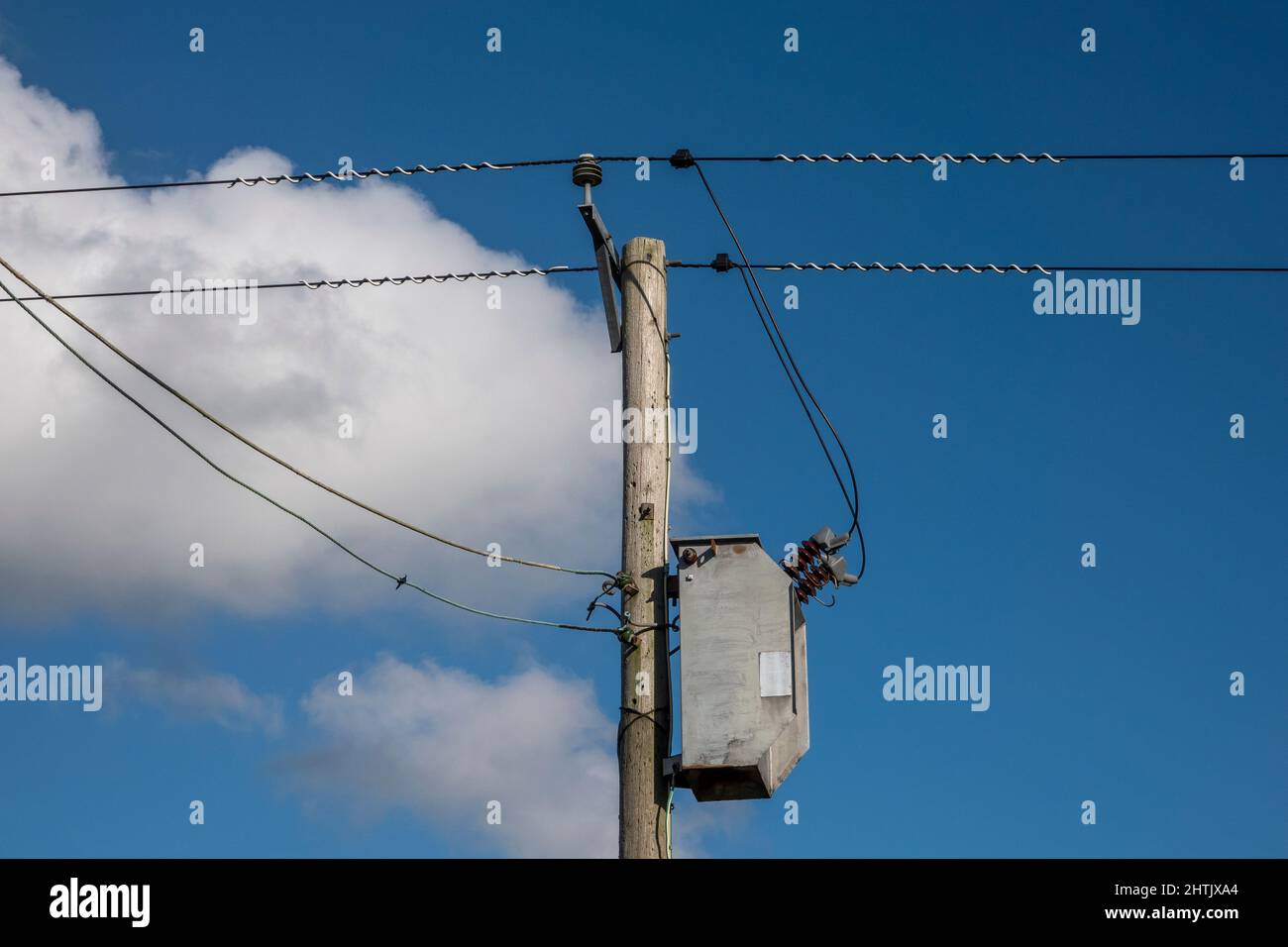 Simple transformer connection on a wooden pylon electricity pole carrying two cables, blue sky and fluffy white cloud behind Stock Photo