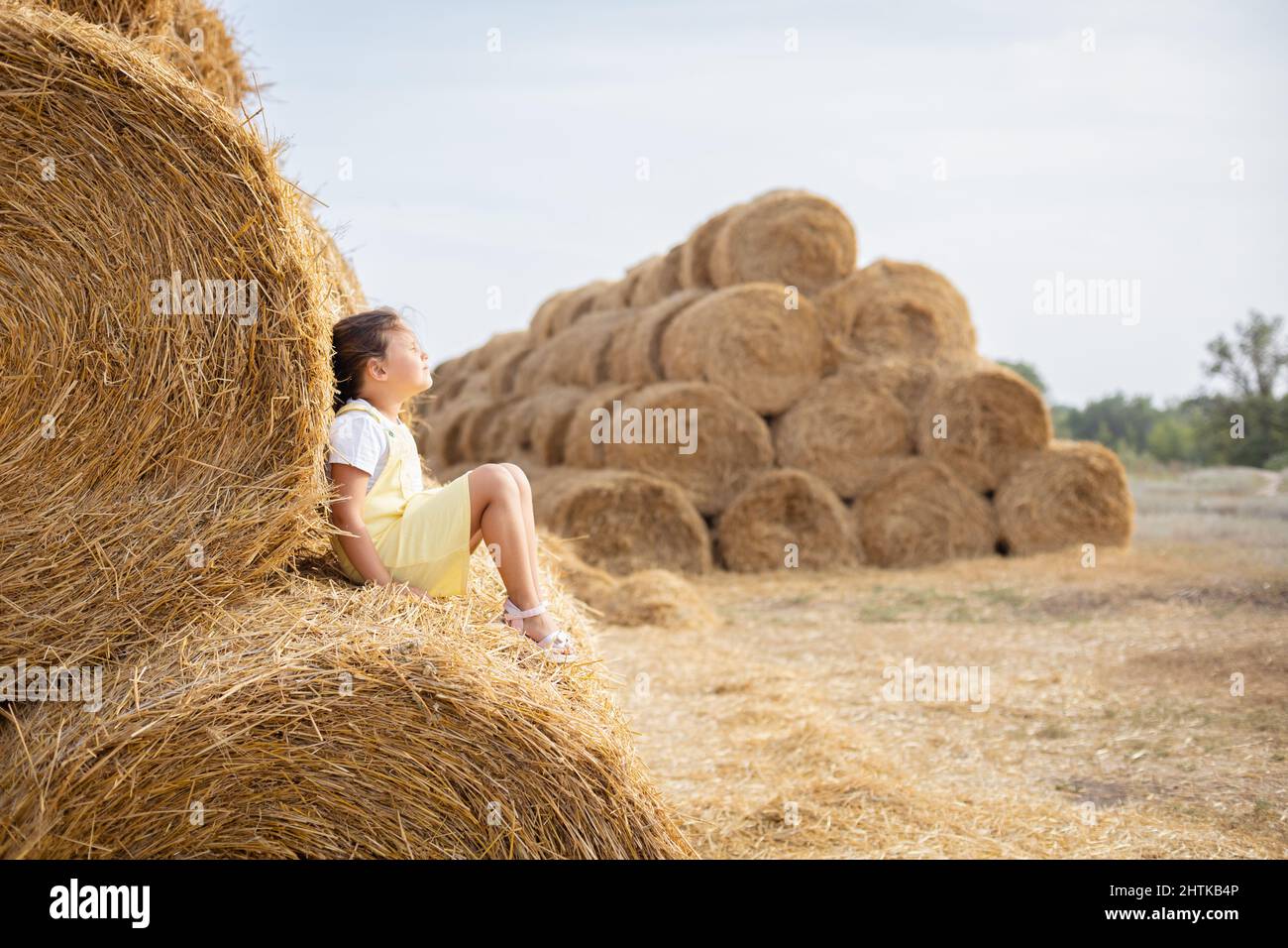 Relaxed young girl seating on high haystack wearing sundress leaning on another hayrick with closed eyes and head up directed in sky. Having fun away Stock Photo