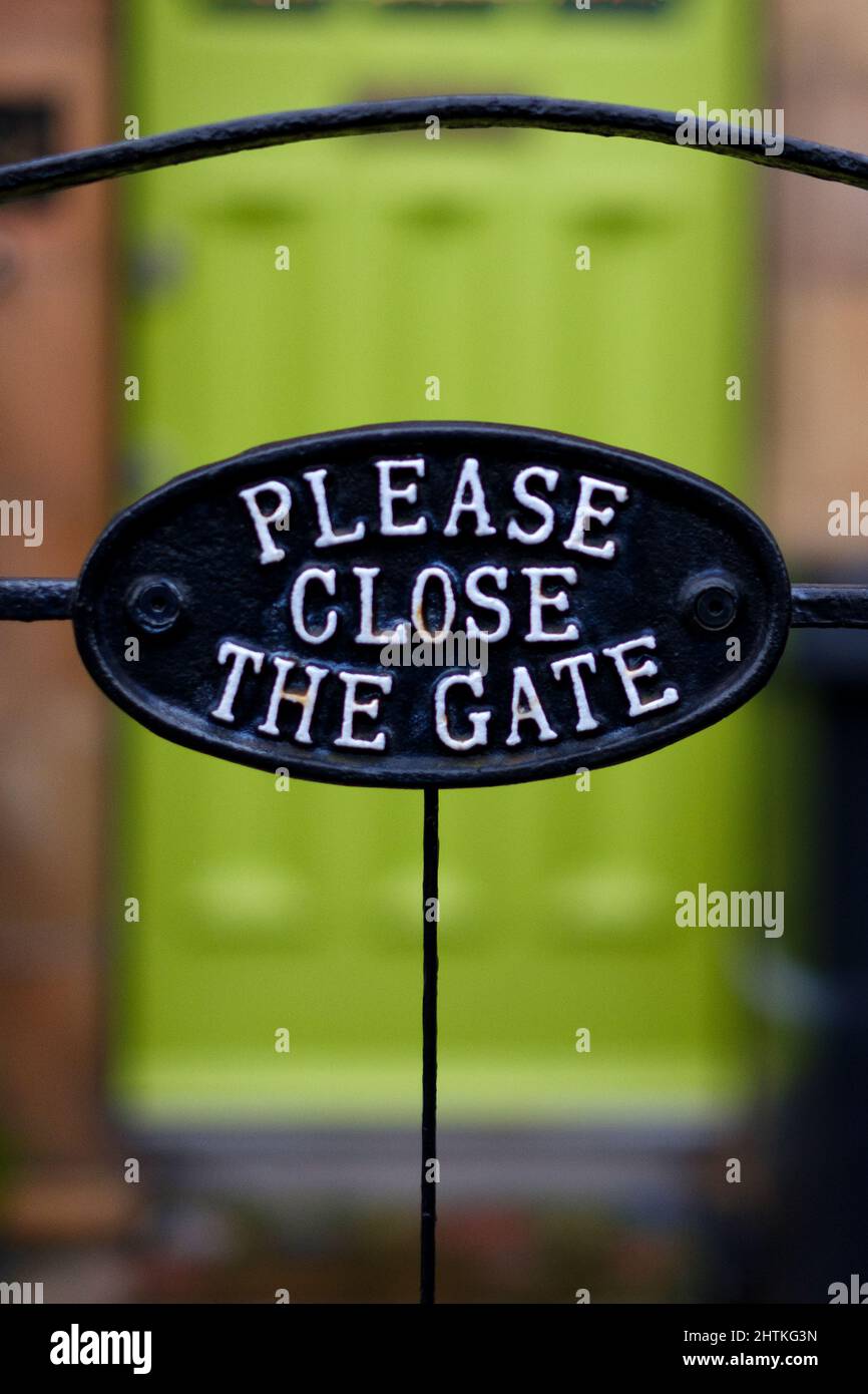 A bold decorative metal cast iron Please Close the Gate sign attached to a metal gate at the end of a garden path to a house with a bright green door. Stock Photo