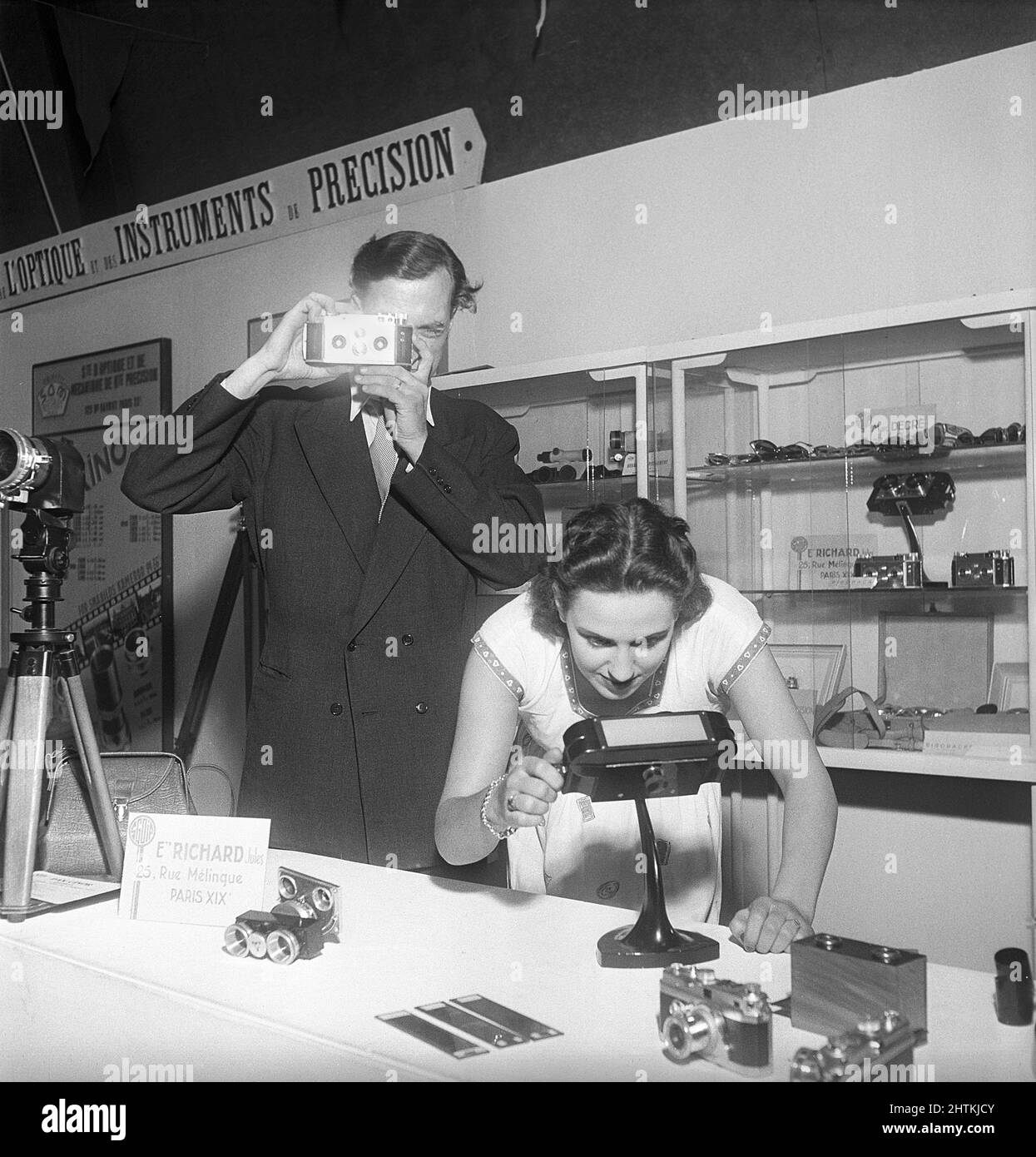 Camera history.  A man with a Jules Richard Paris Verascope holds it up at a trade fair.  The camera took twin shots of the scene, and the slides could then be viewed in dedicated image viewers to experience the depth and effect of the twin lense photography. The woman demonstrates such a viewer that is visible on the table. Sweden 1951 Kristoffersson ref BD95-12 Stock Photo
