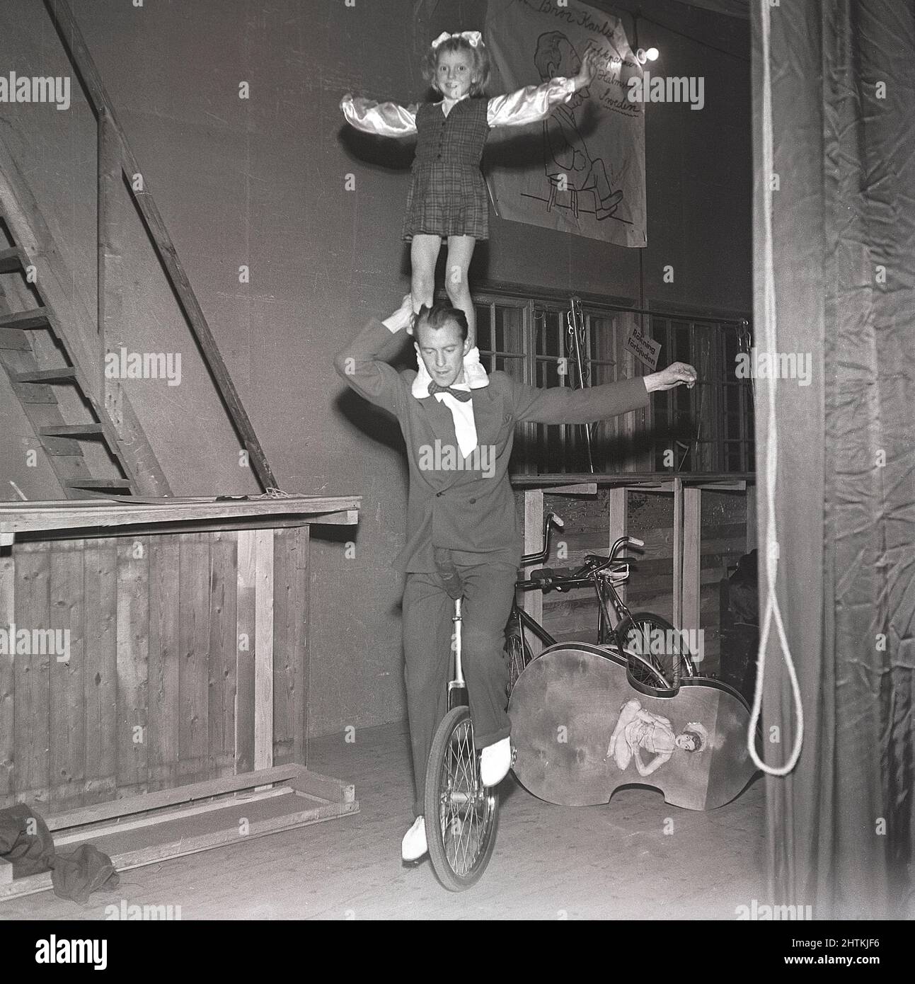 1950s bicycle performer. A man is balancing a girl on his shoulders while cycling around on a theatre stage on a unicycle. Sweden 1951 Kristoffersson ref BE61-6 Stock Photo