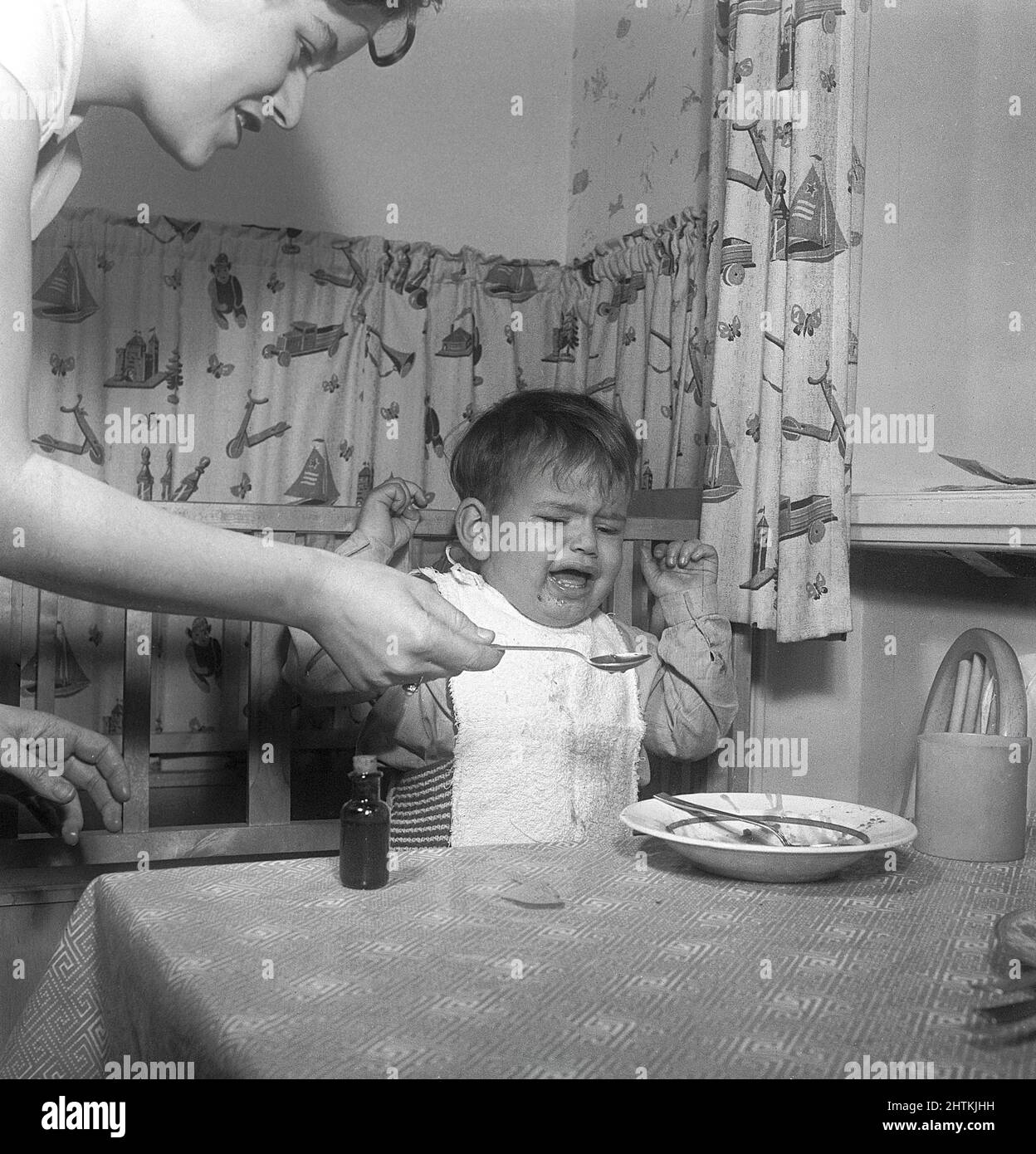 Boy in the 1950s. An angry little boy is being spoon fed his food against his will. He looks upset and angry and is not willing to eat more. The still positive and smiling mother does her best to make him. Sweden 1951 Kristoffersson Stock Photo