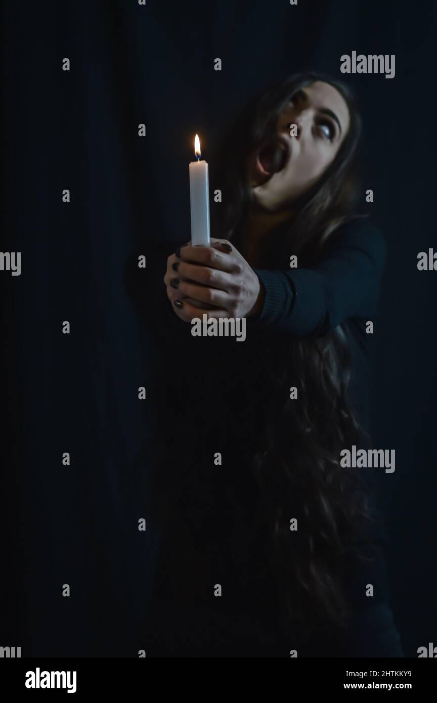 Creepy portrait of a young woman with very long hair who is holding a lit candle and staring backwards with white eyes and mouth open Stock Photo