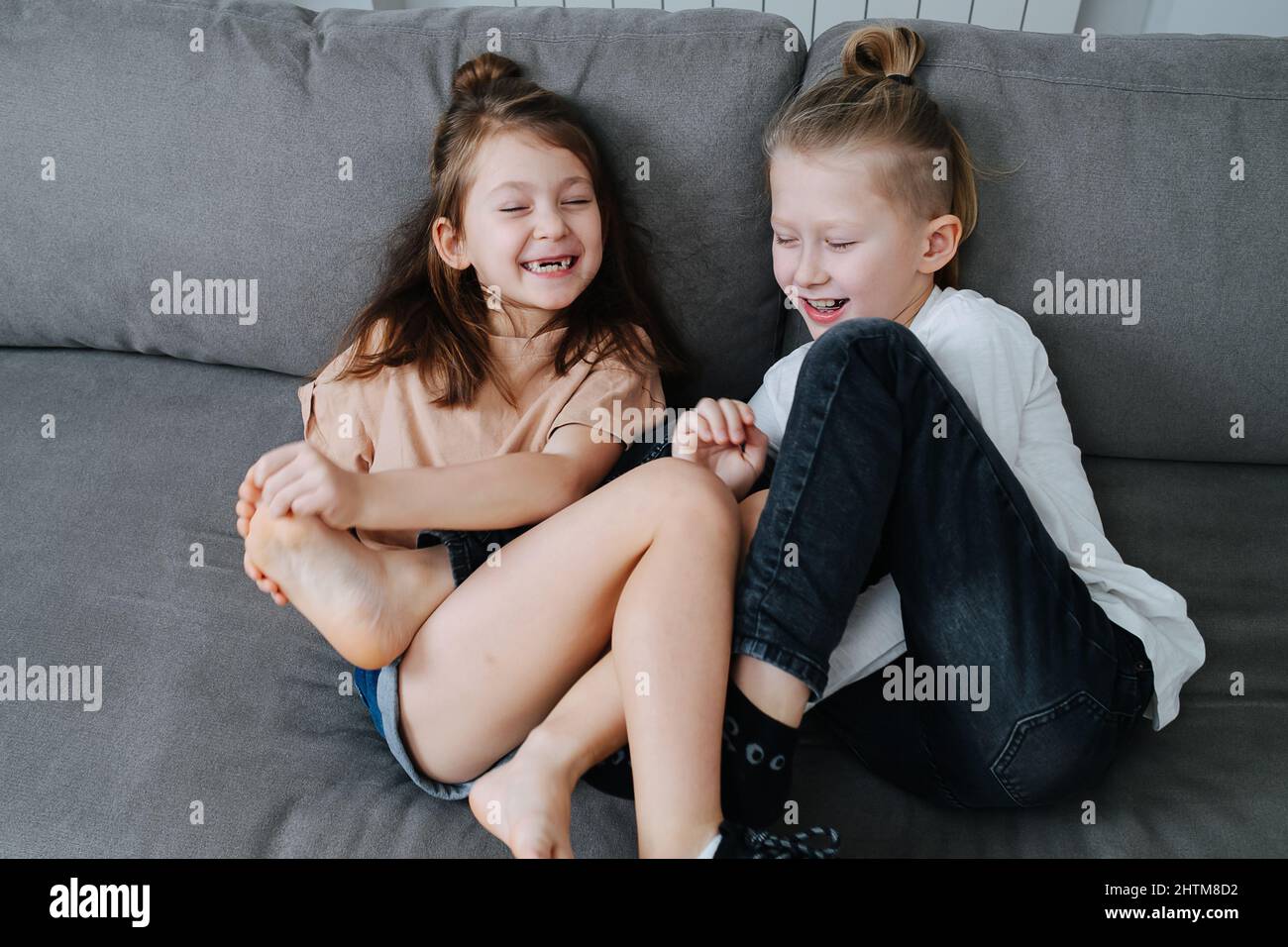 Two kids having a good time, trying to tickle each other's feet. They sit on a couch, eyes closed, having a laugh. Stock Photo