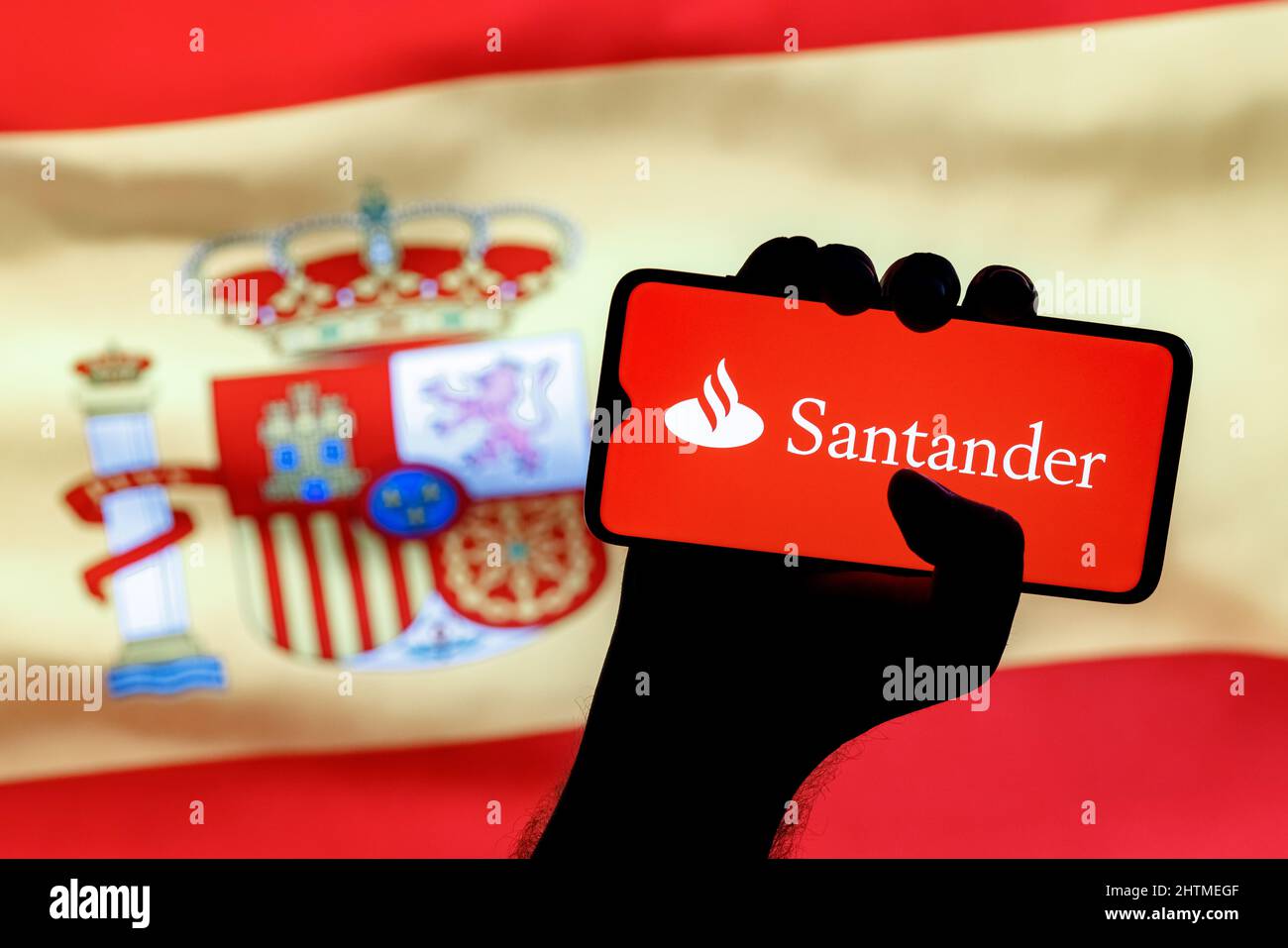 Smartphone with Santander bank logo in clenched hand on background of Spain flag Stock Photo