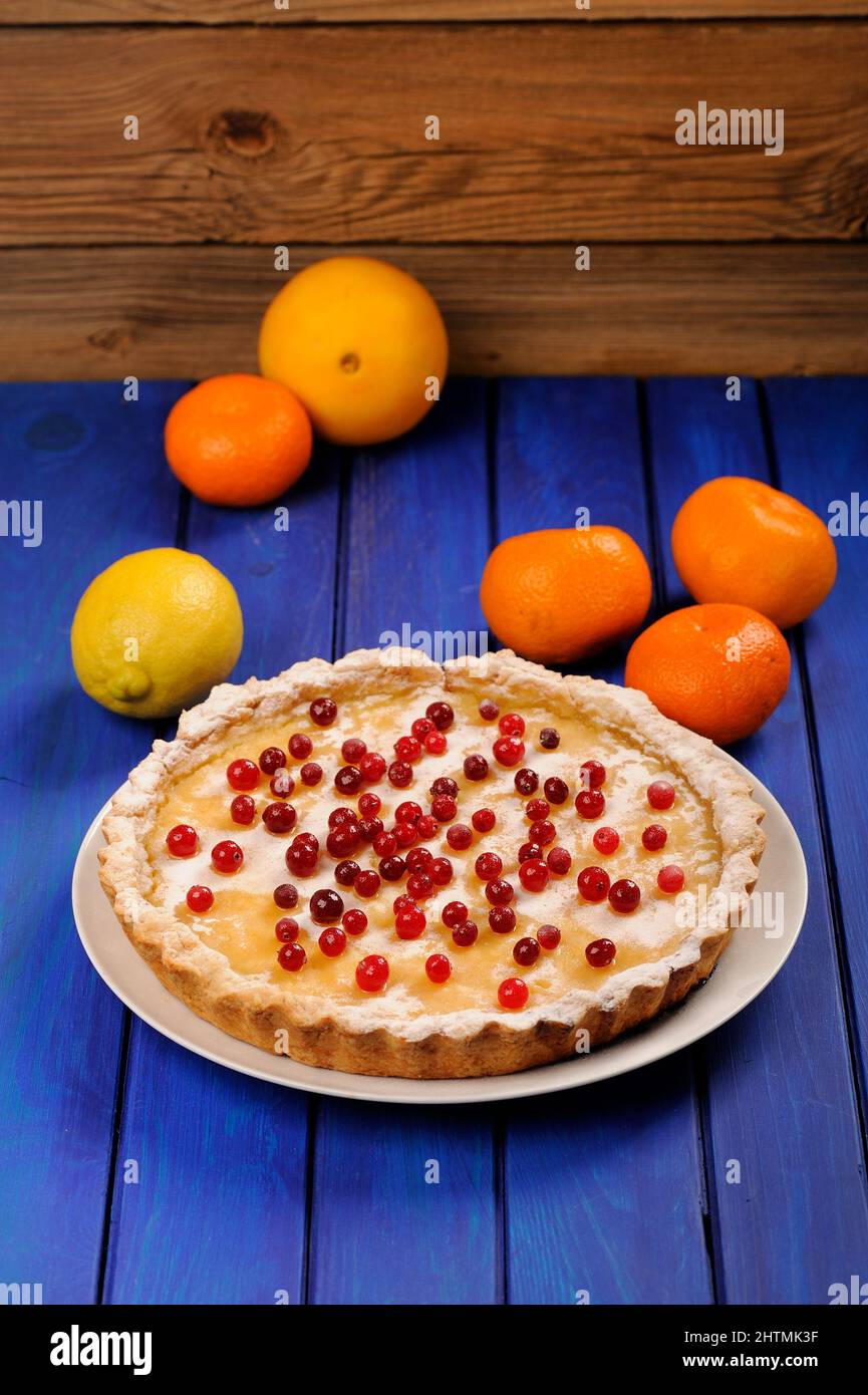 Homemade round lemon pie decorated with fresh cranberries and whole orange, lemon and clementines on deep blue table  vertical Stock Photo