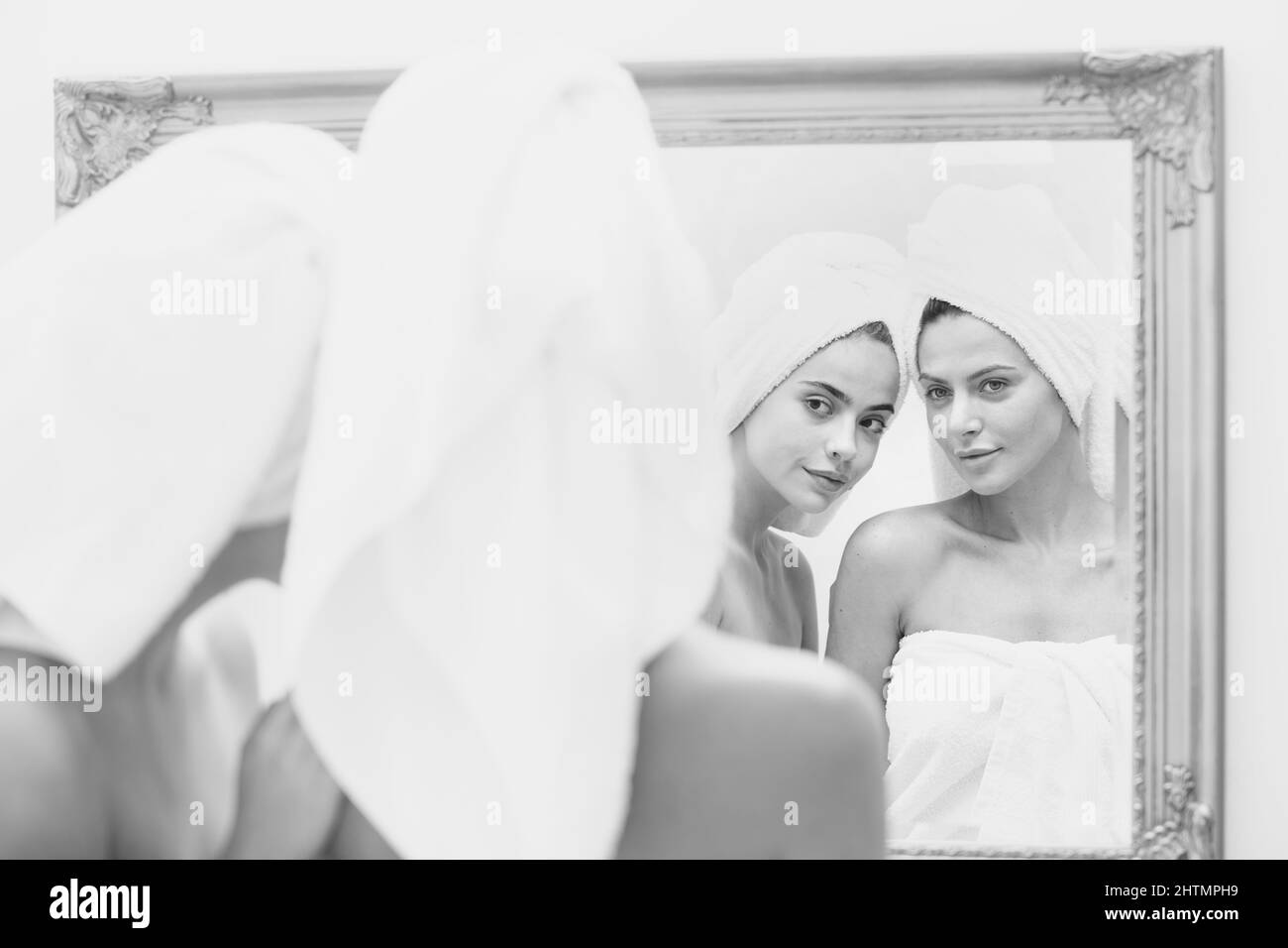 Closeup beautiful woman look in mirror after shower. Young pretty girls friends wrapped in towel smoothing perfecting skin, daily morning routine Stock Photo