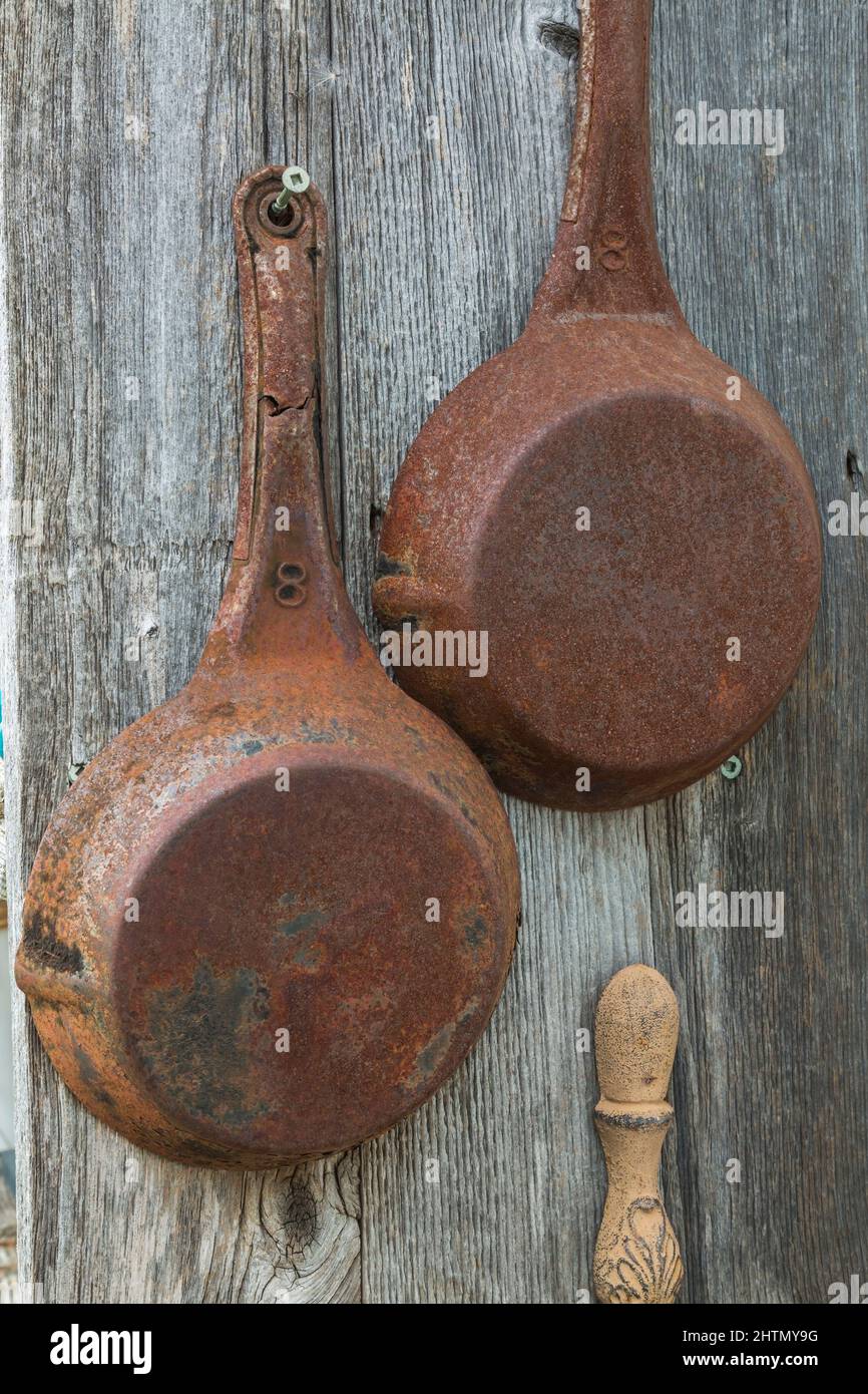 Old rusted frying pans hanging on grey weathered exterior wall of old rustic wood plank cabin. Stock Photo