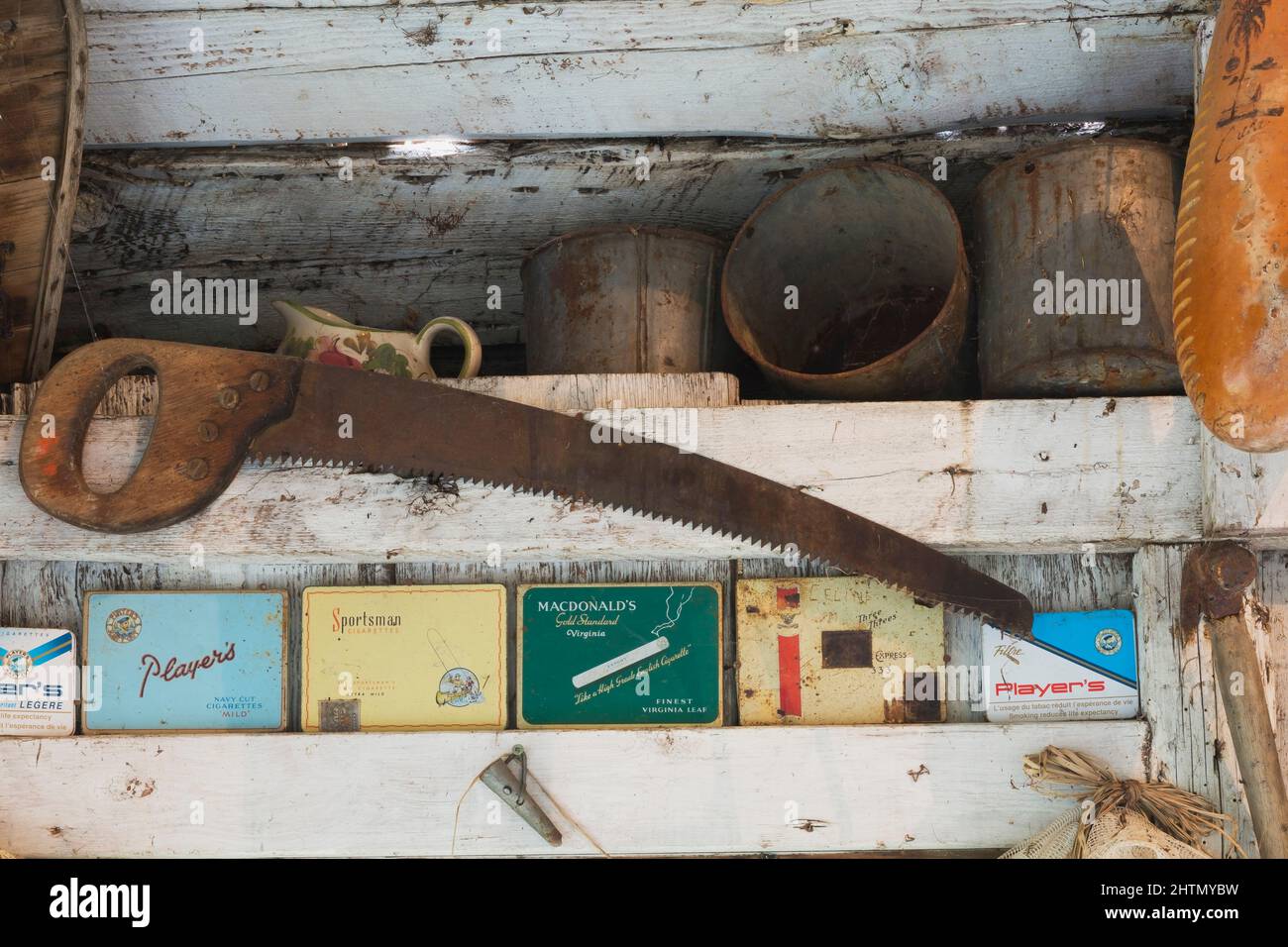 Antique saw, empty paint cans, metal cigarette boxes on shelf inside old rustic wood plank cabin. Stock Photo