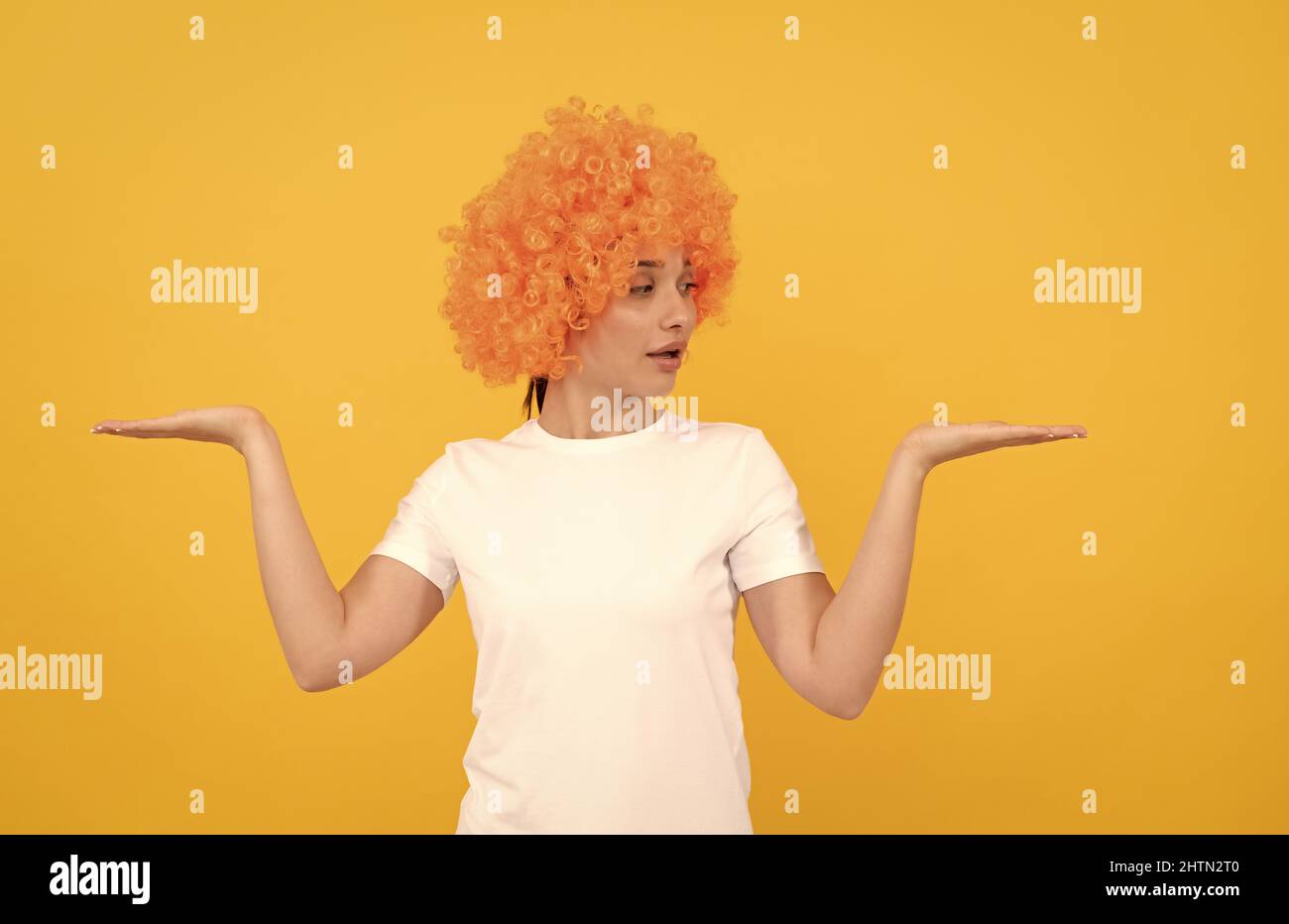 amazed funny girl with fancy look wearing orange hair wig presenting product, copy space, choice. Stock Photo