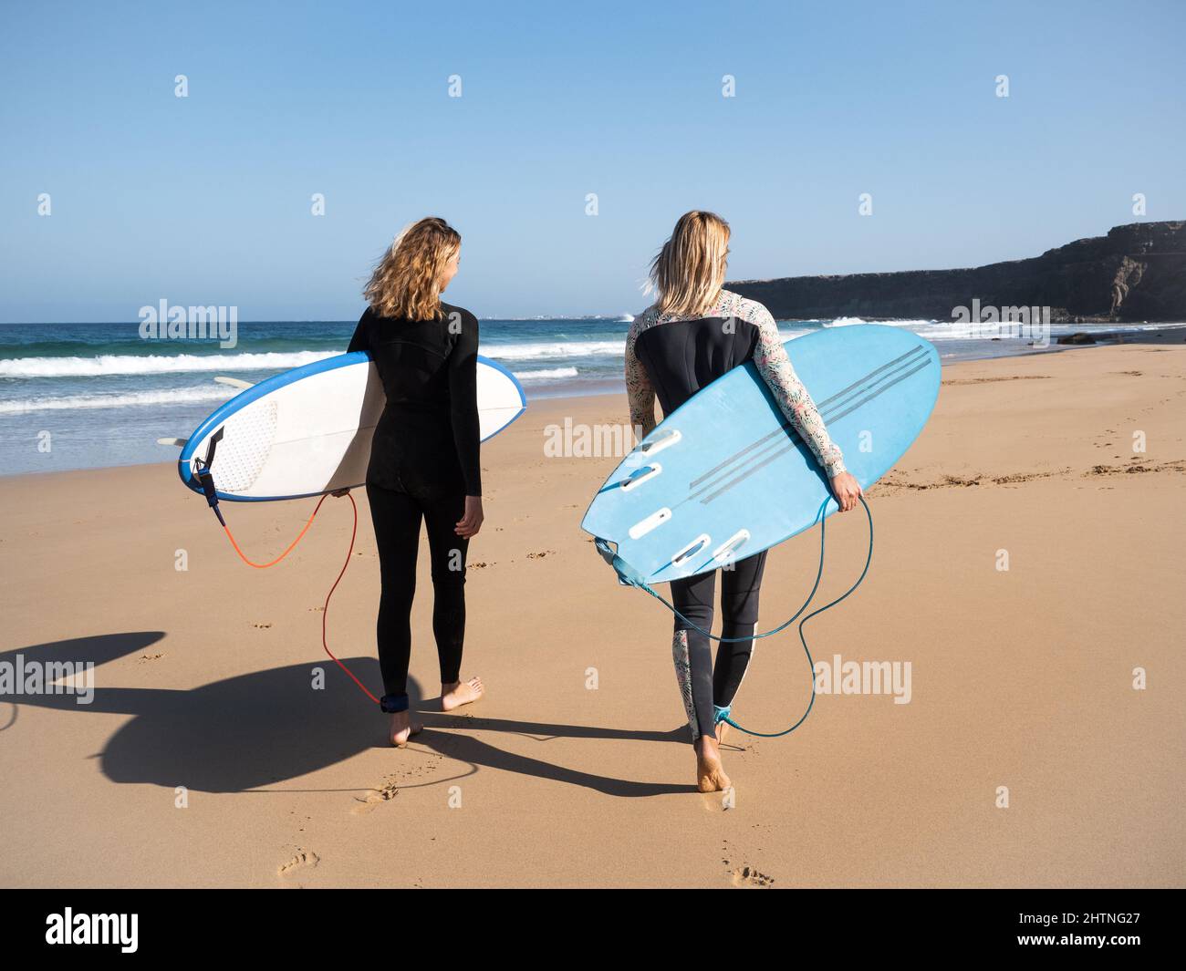 2 caucasian surfer women on the beach walking to the waves. They are wearing winter wetsuits. Stock Photo