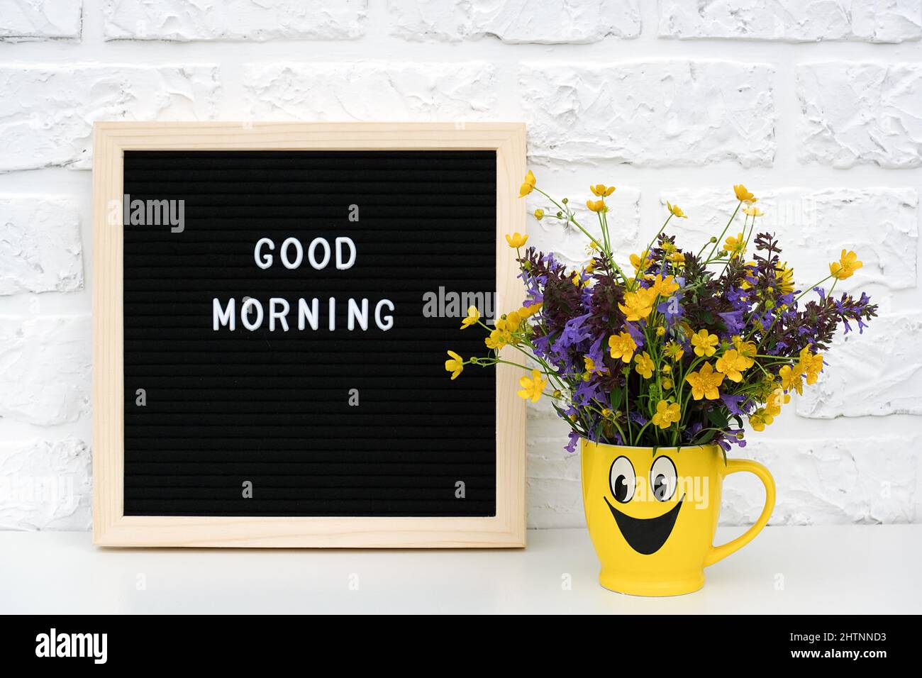 Text Good morning on black letter board and bouquet of colored flowers in yellow cup with painted funny face on table against white brick wall. Creati Stock Photo