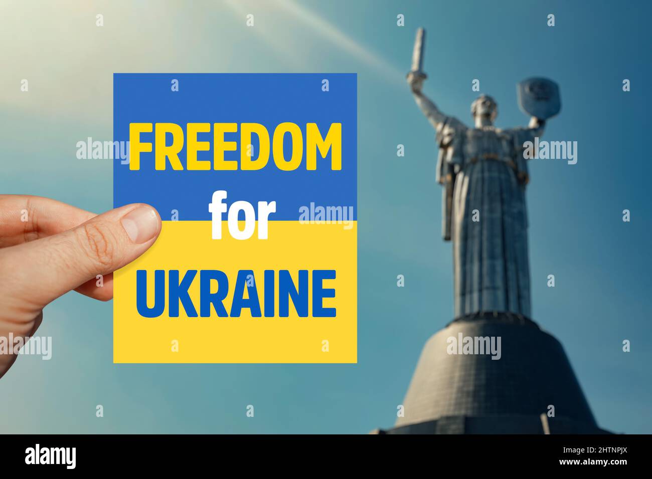 Male hand holding Ukraine flag card with Freedom for Ukraine  message in front of blurry Mother Land monument in Kiev. Stock Photo