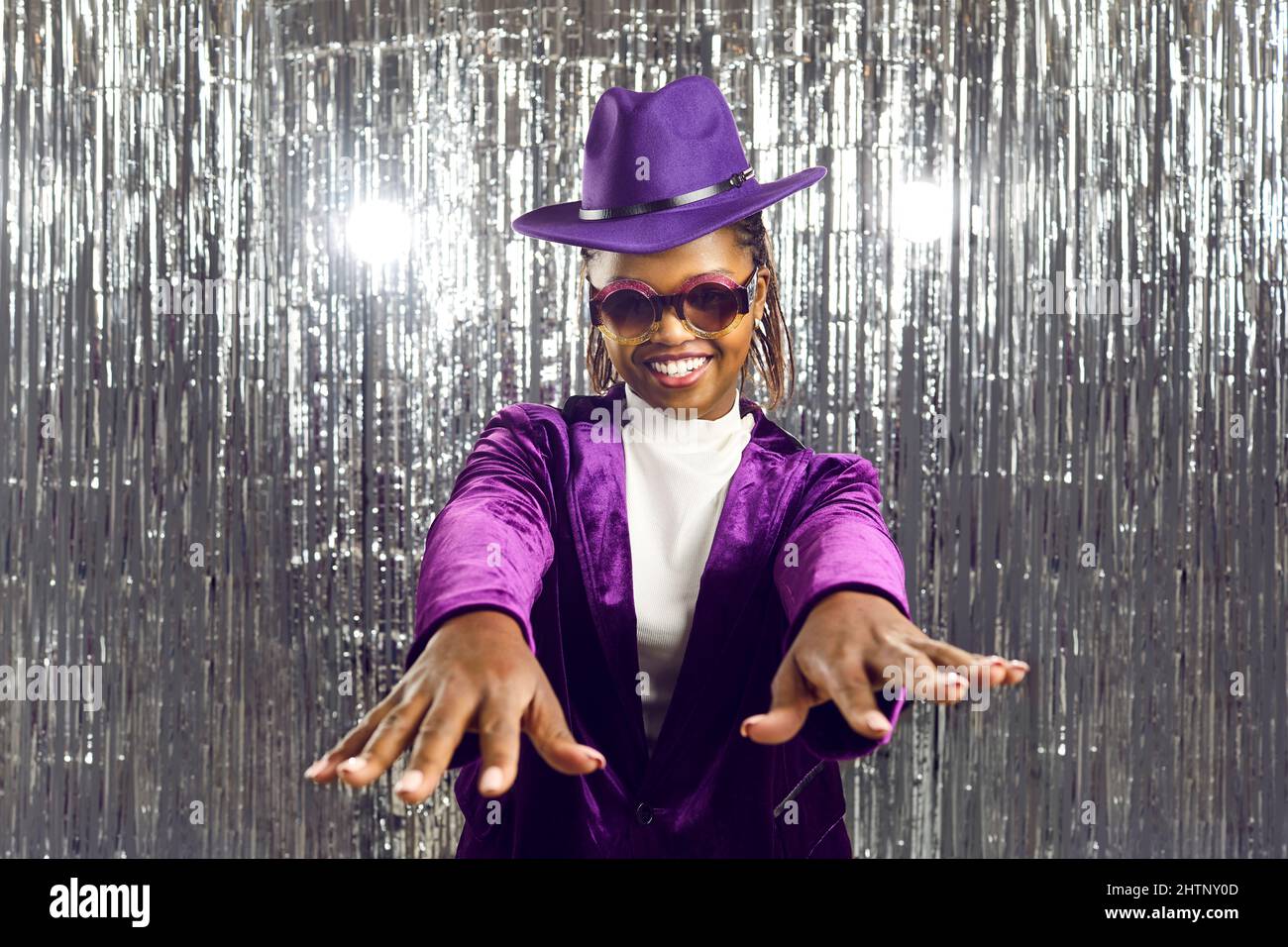 Happy young African American woman in a purple hat and disco glasses dancing at a party Stock Photo