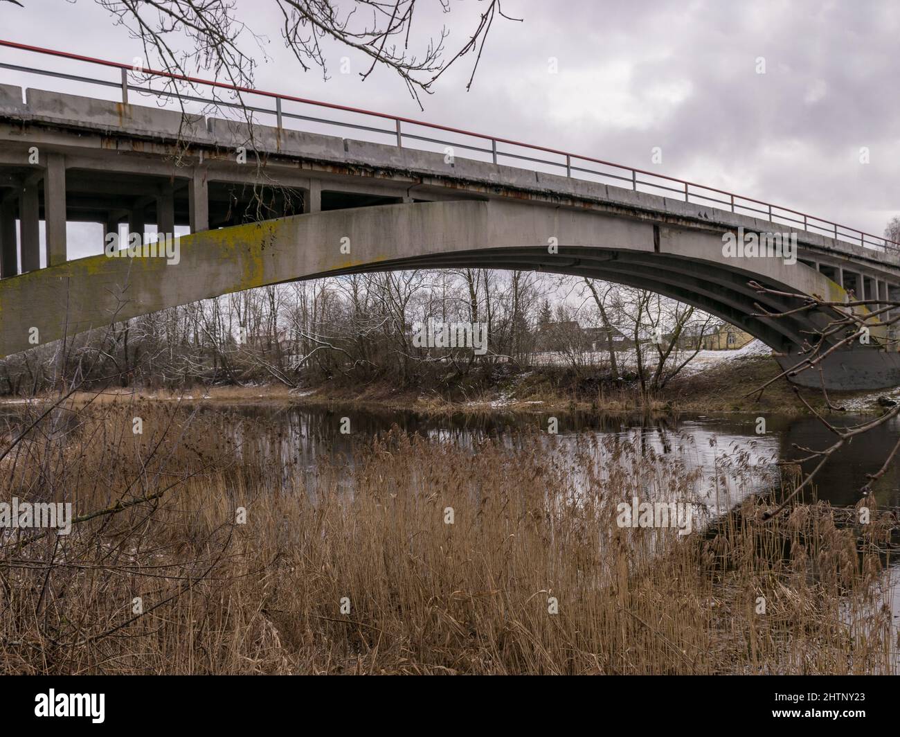 concrete arched bridge over a small and wild river, overgrown with shrubs, withered branches and old grass Stock Photo