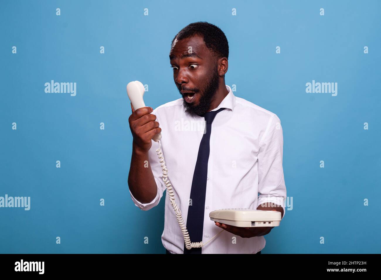 Shocked businessman looking in awe at reciever of cord telephone after losing argument with person shouting at the other end of the line. Office worker holding phone cannot belive what he hears. Stock Photo