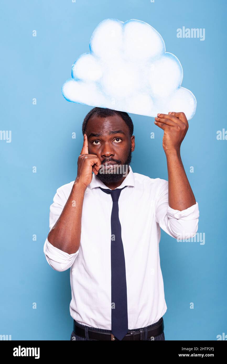 Portrait of professional businessman pointing at white paper idea cloud standing in front of blue background. African american entrepreneur thinking under thought bubble. Stock Photo