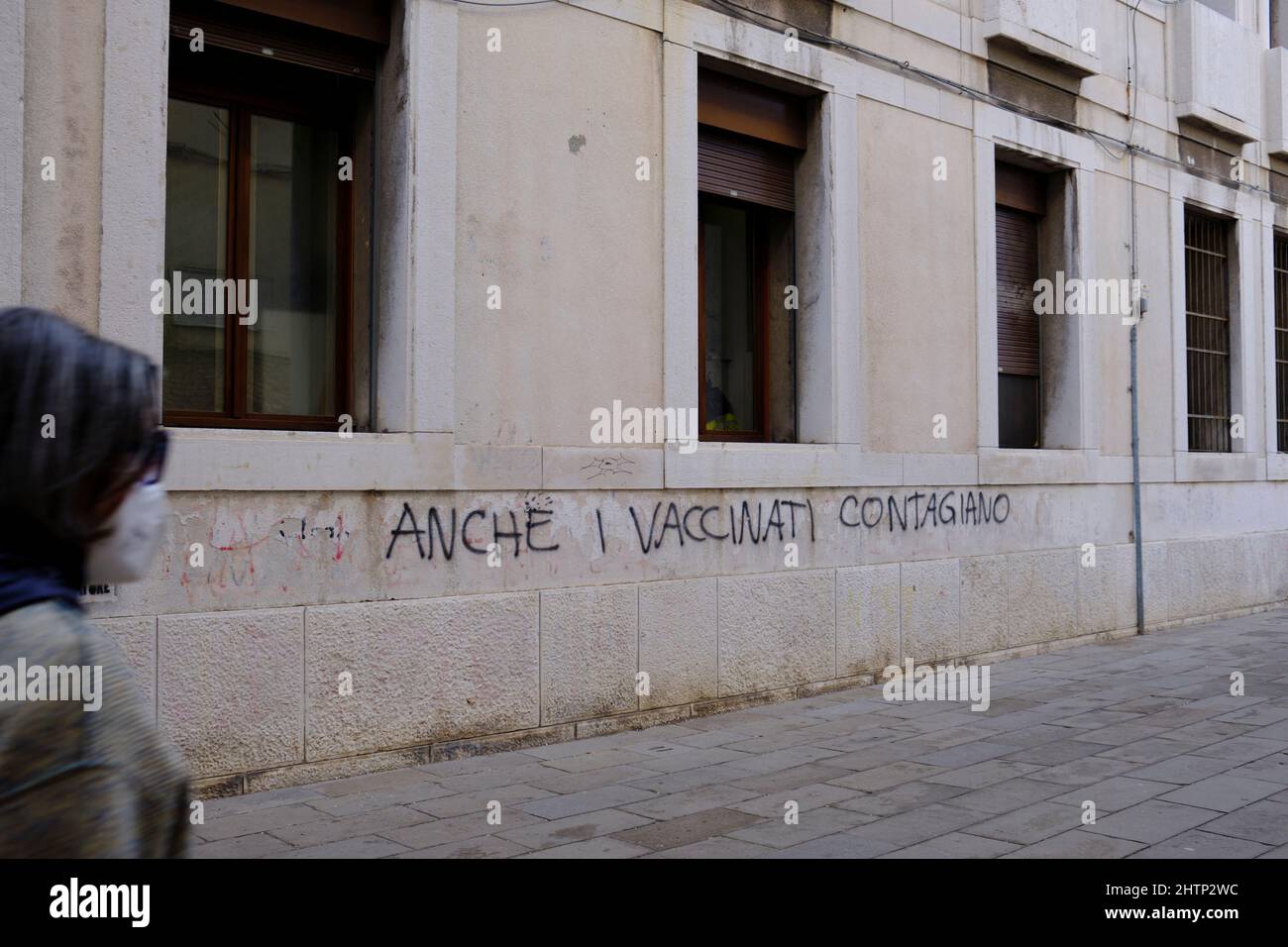 woman wearing mask walks past anti covid graffiti in Venice (even the vaccinated are contagious) Stock Photo