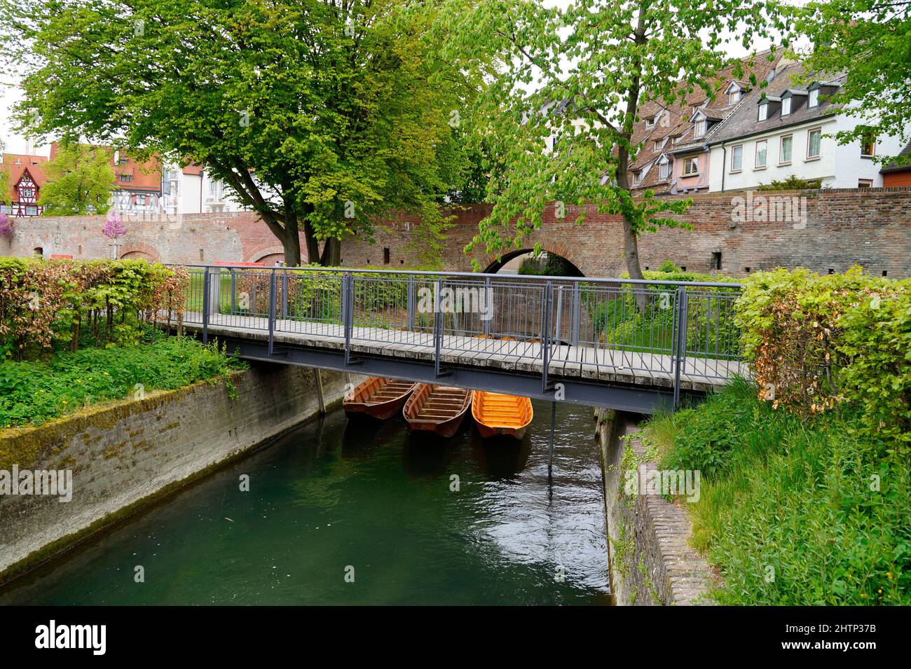a scenic view of a little bridge, wooden boats (Ulmer Schachteln) underneath it, green trees and the old town wall in the city of Ulm in Germany Stock Photo