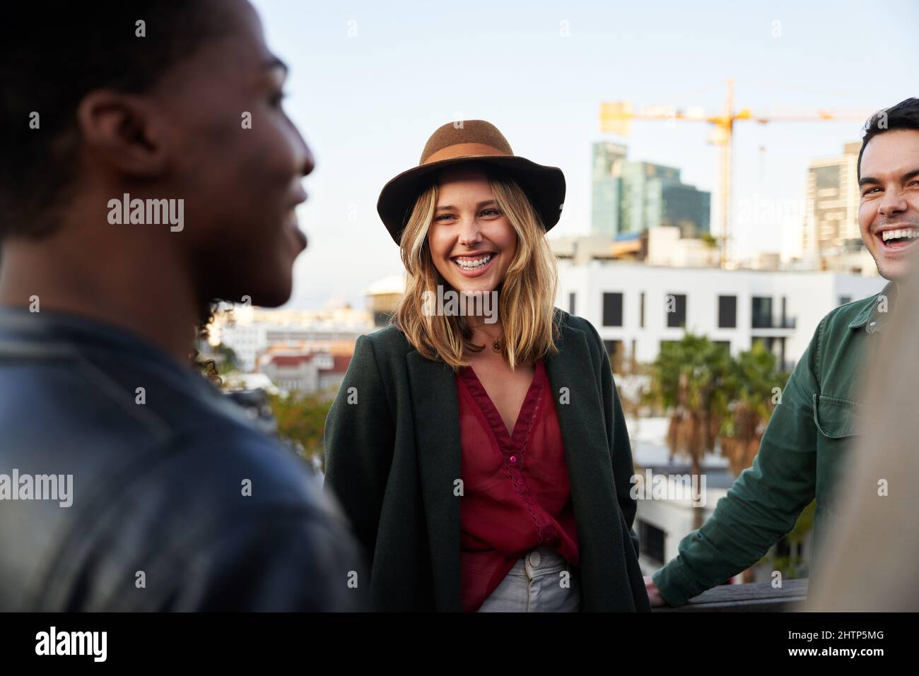 Caucasian female smiling with group of multi-cultural friends socializing on a rooftop terrace at dusk. Stock Photo