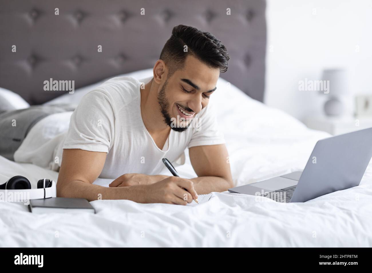 Distance Learning Concept. Millennial Arab Guy Using Laptop While Lying In Bed Stock Photo