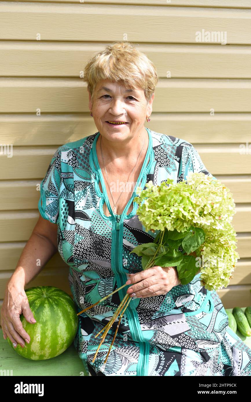 Portrait of 69 y.o. woman sitting on a bench and holding watermelon and bouquet of hydrangea Stock Photo
