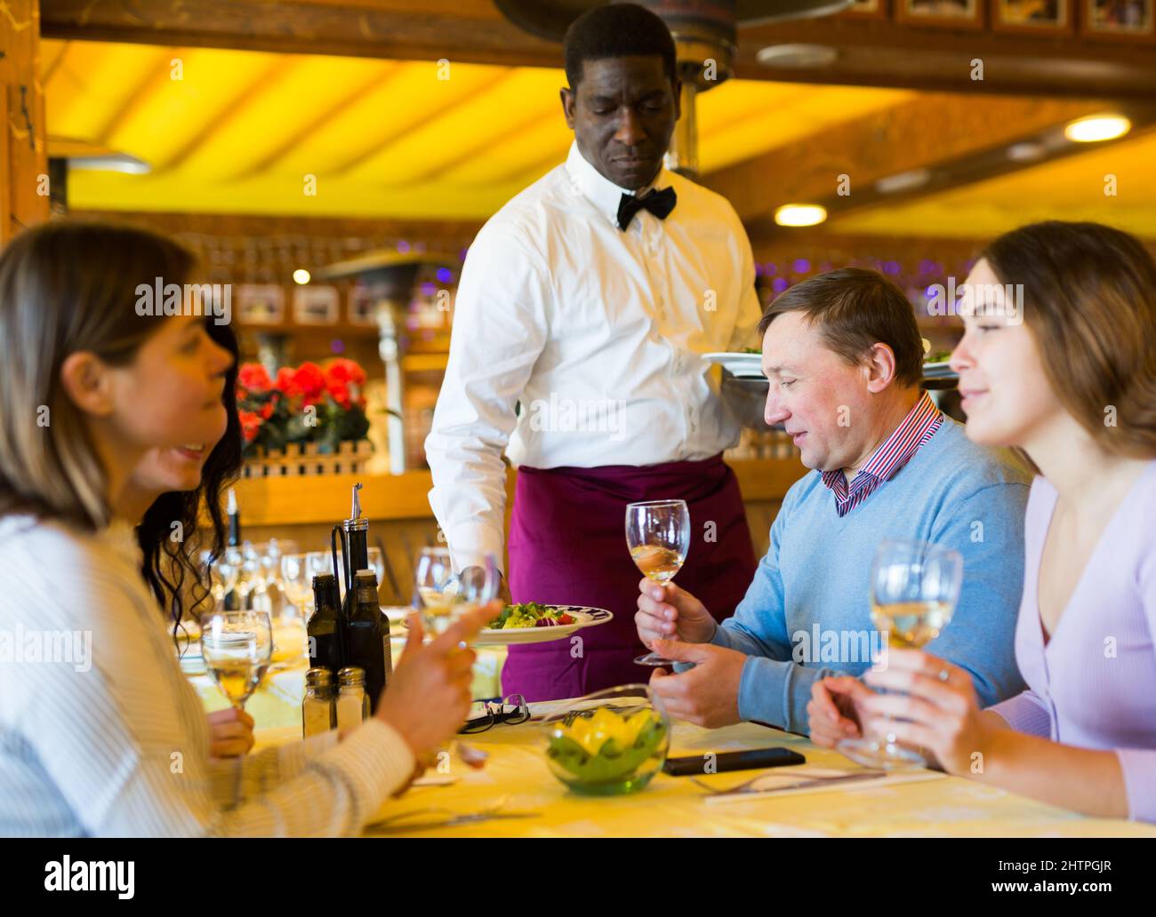 African american waiter serving dishes to smiling adults having dinner in restaurant Stock Photo